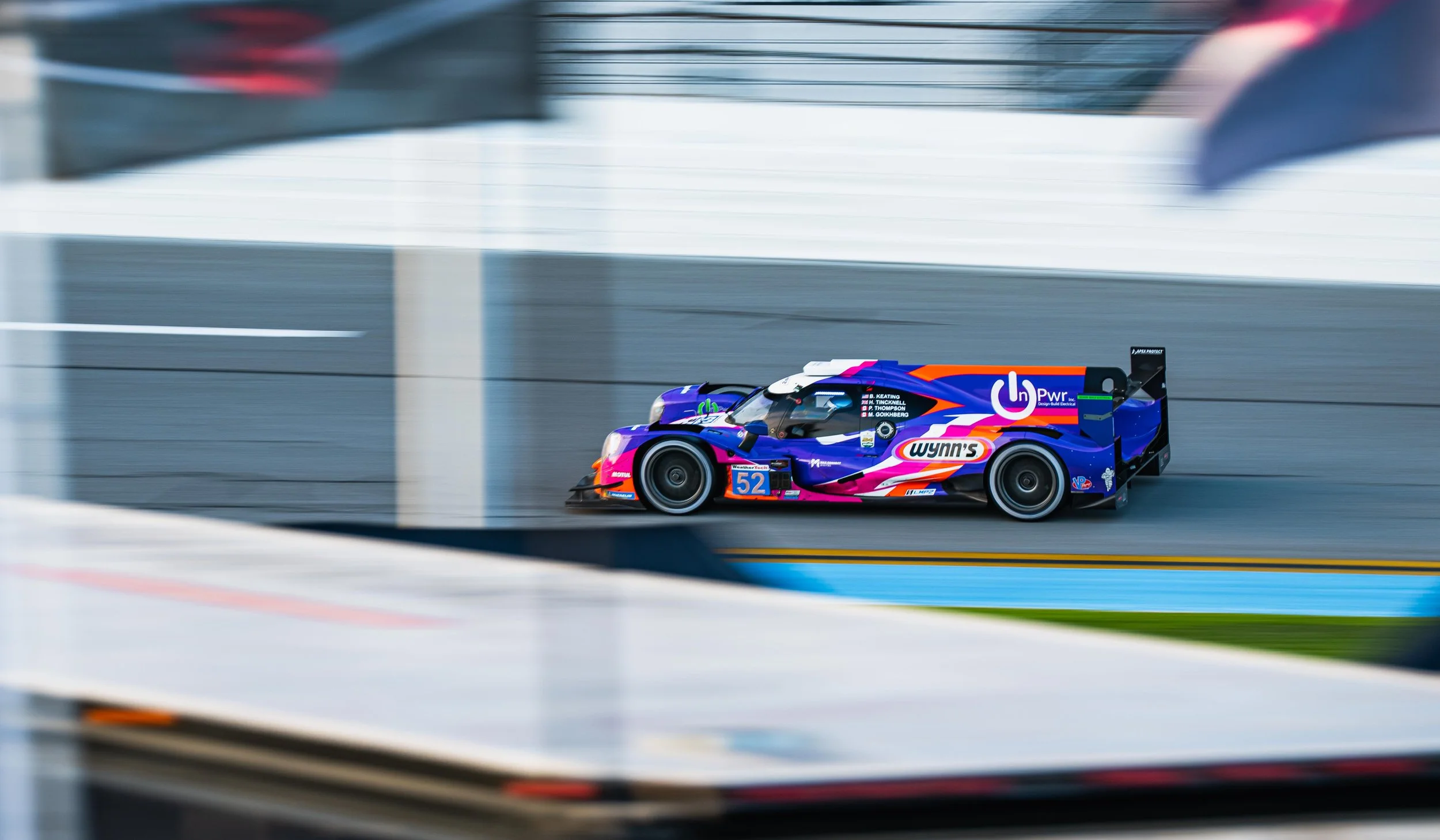 A colorful race car with number 52 speeding on a race track, seen through a blurred barrier.