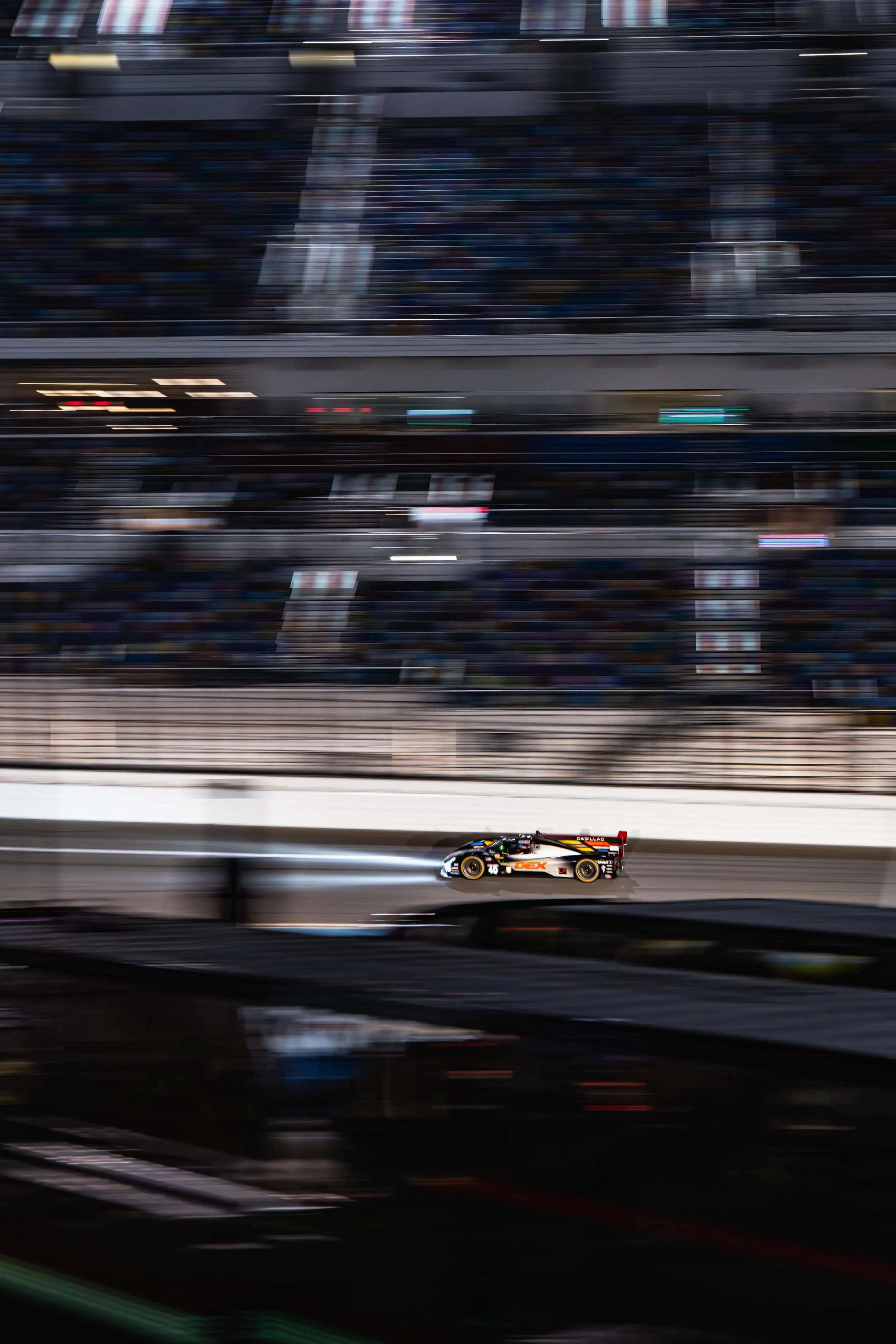 A race car moving on a track at night, with blurred grandstands and spectators in the background.