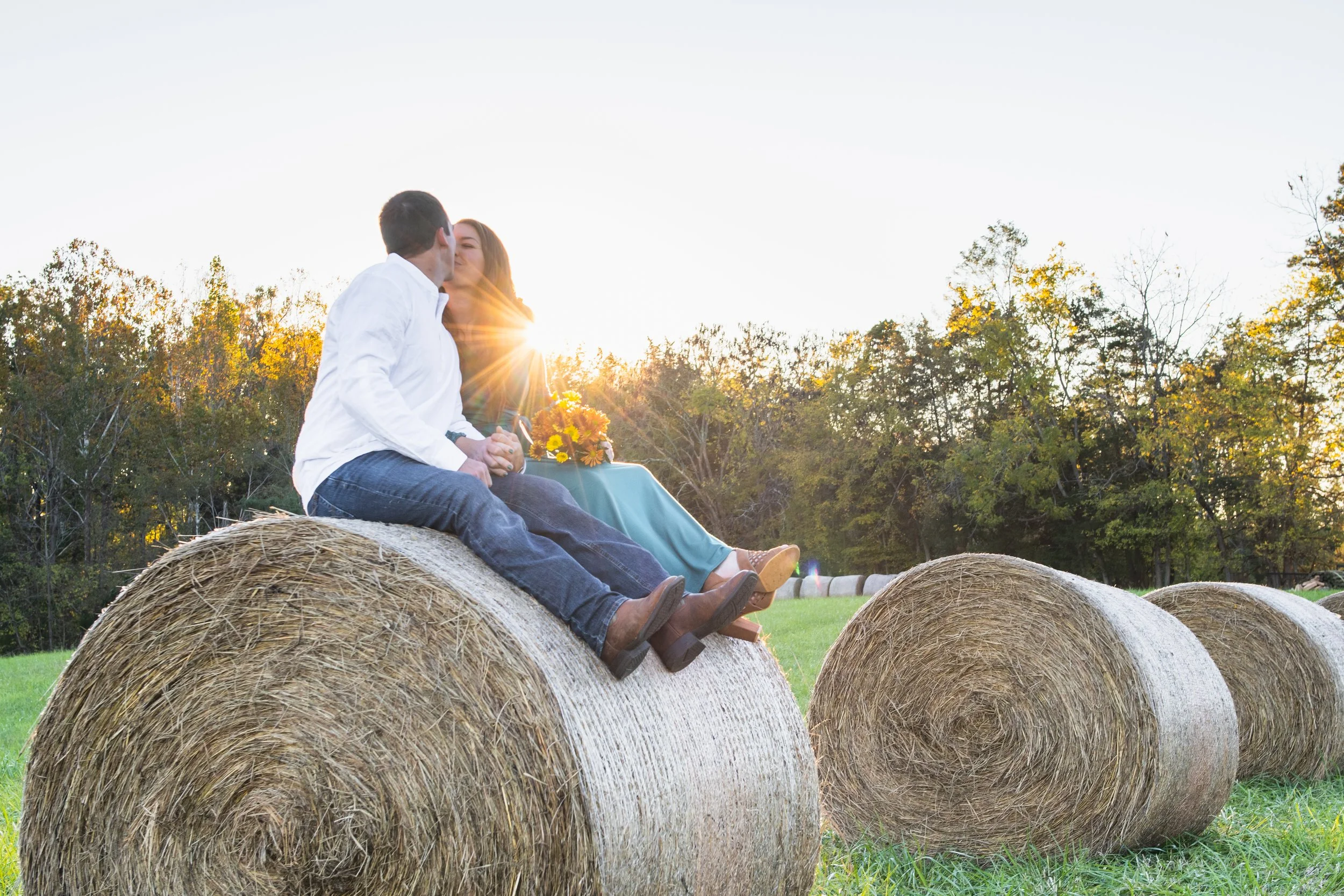 A couple sitting on hay bales in a field at sunset, kissing with the sun shining behind them.