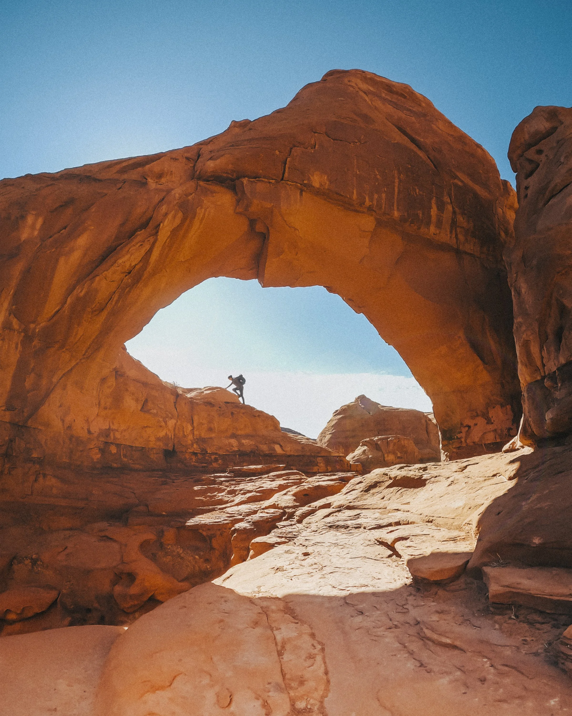 A hiker climbing on orange rock formations near a natural stone arch under a blue sky.