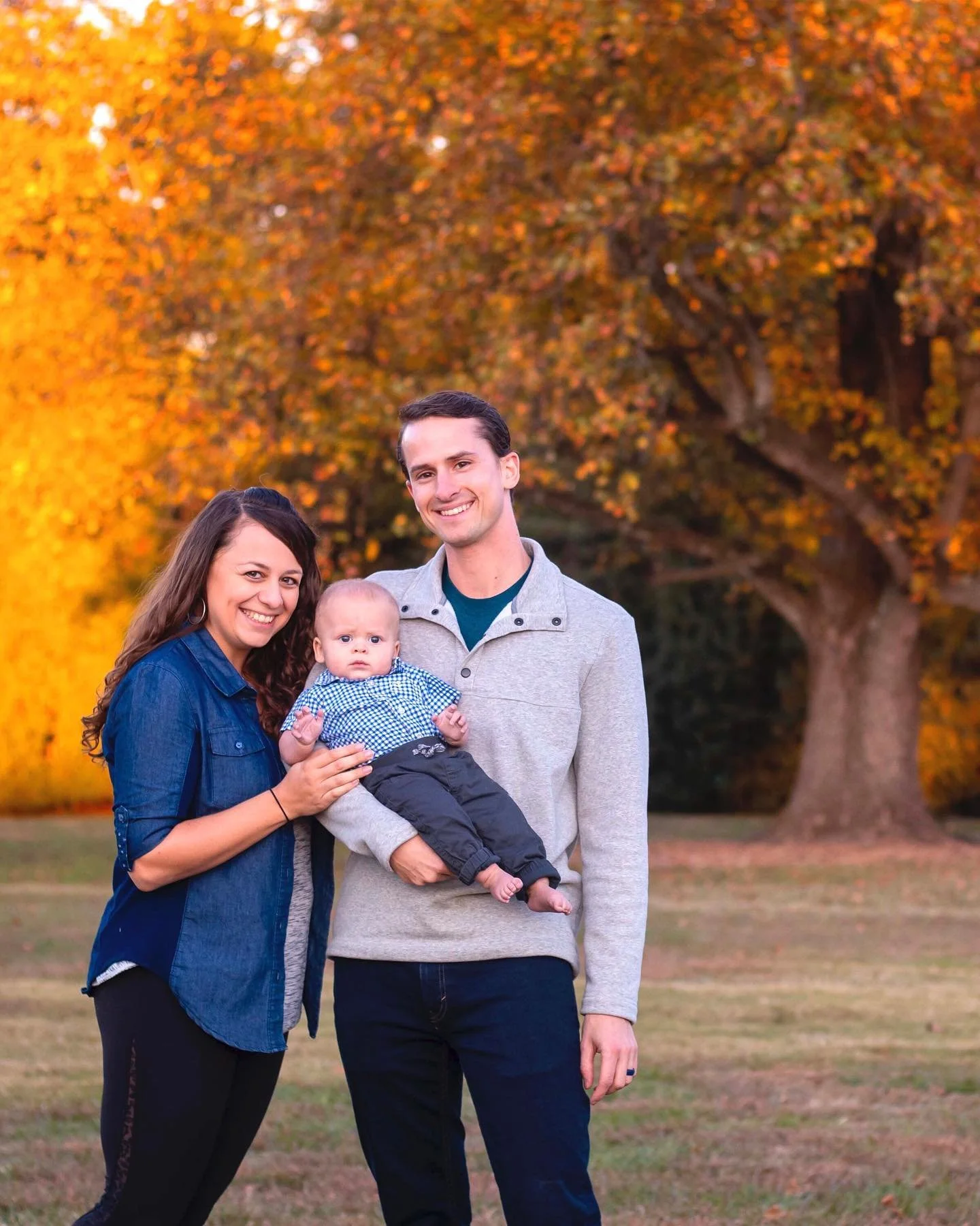 A smiling family of three standing outside in fall with orange and yellow trees in the background. The mother holds a baby boy in front of a father who is dressed in a light gray sweater.