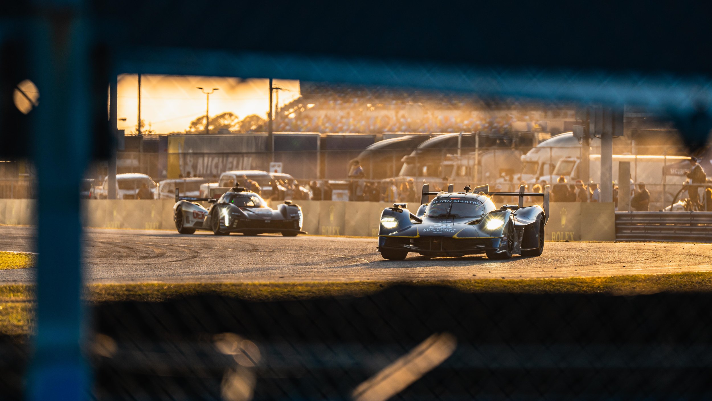 Race cars on a track during sunset, viewed through a fence with spectators and trucks in the background.