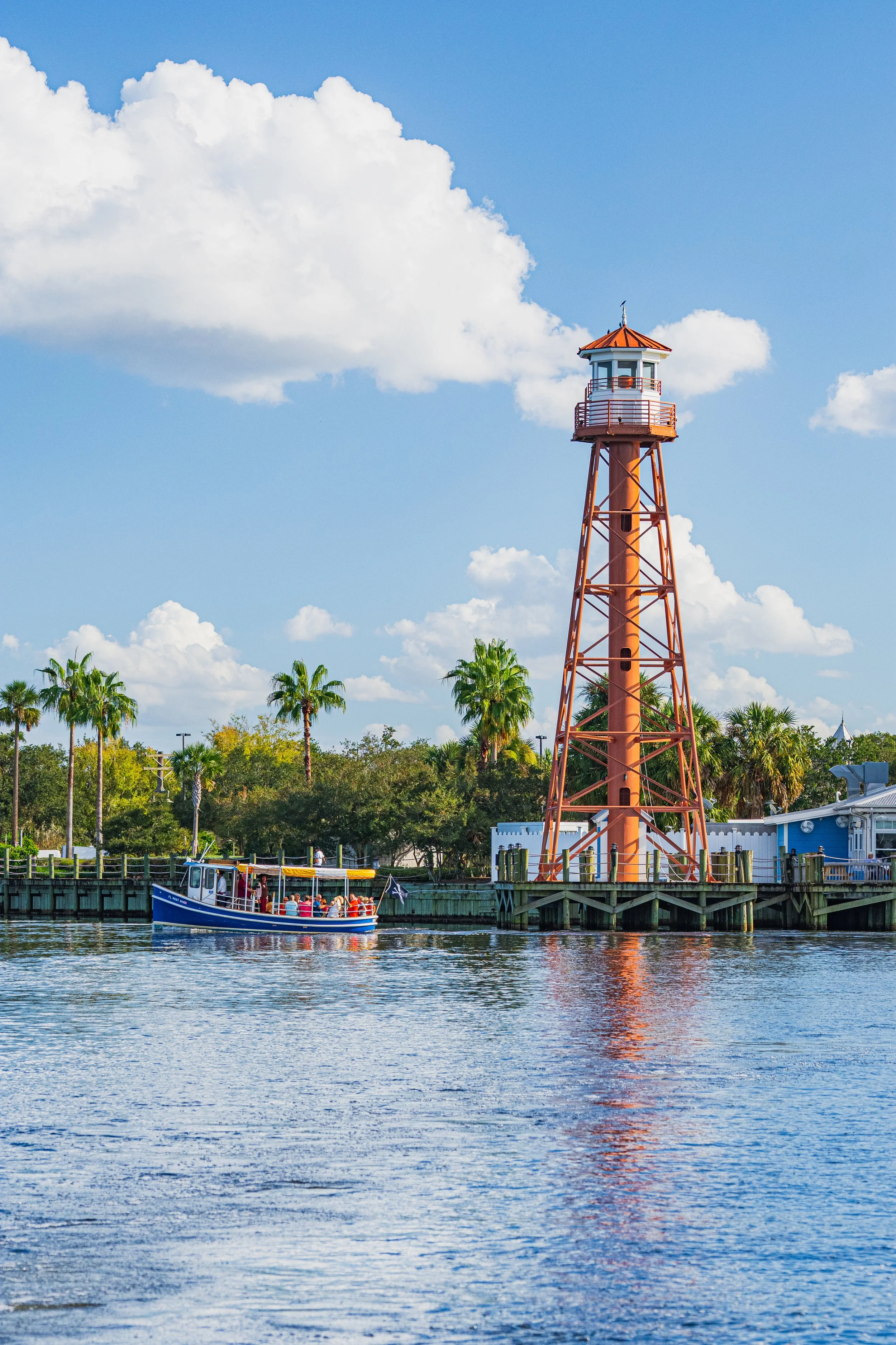 A waterfront scene with a red-orange lighthouse, palm trees, a boat with people, and a partly cloudy sky. 