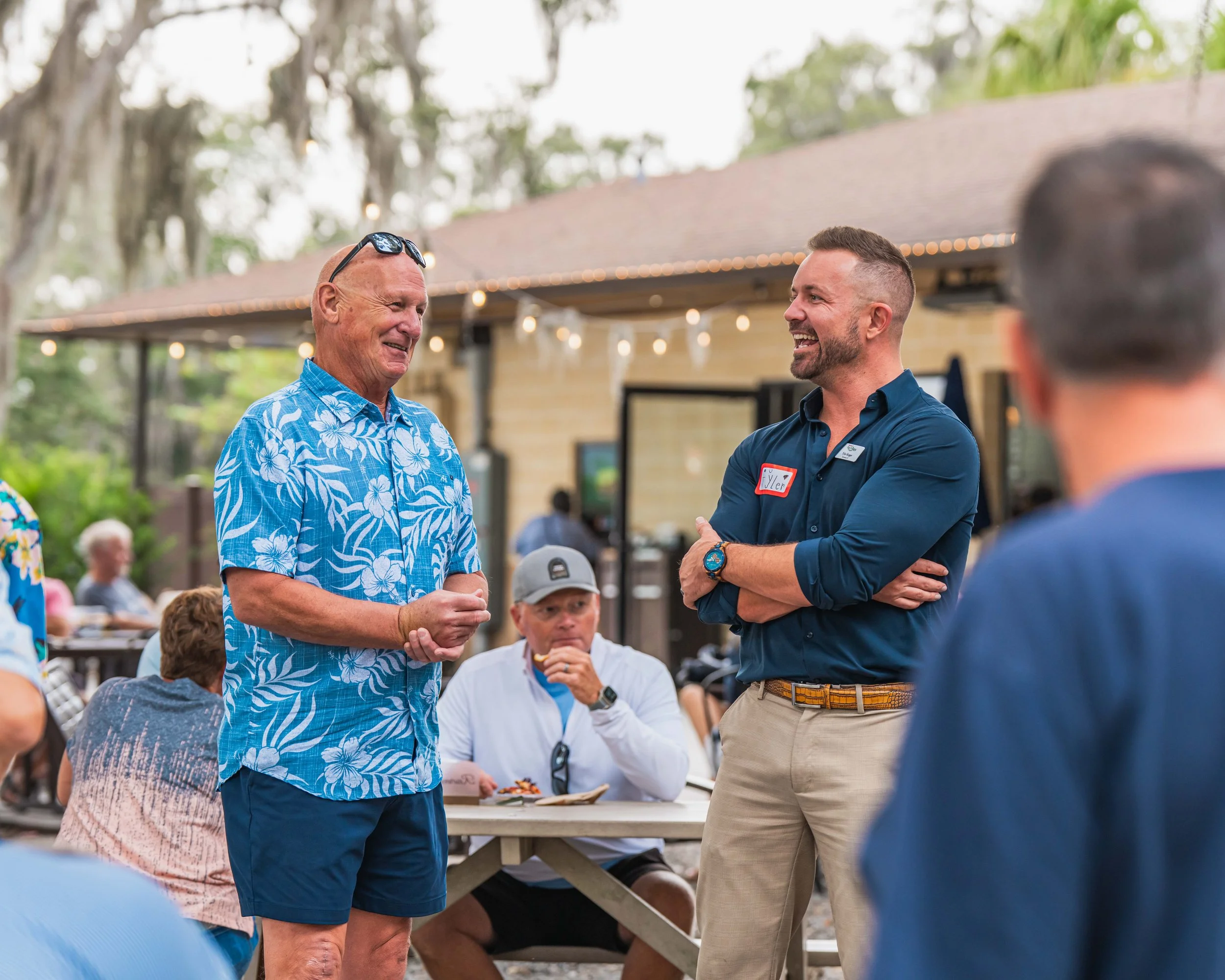 Two men are smiling and talking at an outdoor gathering, one wearing a blue Hawaiian shirt and the other in a dark blue dress shirt with a red name tag, with people seated at picnic tables in the background.
