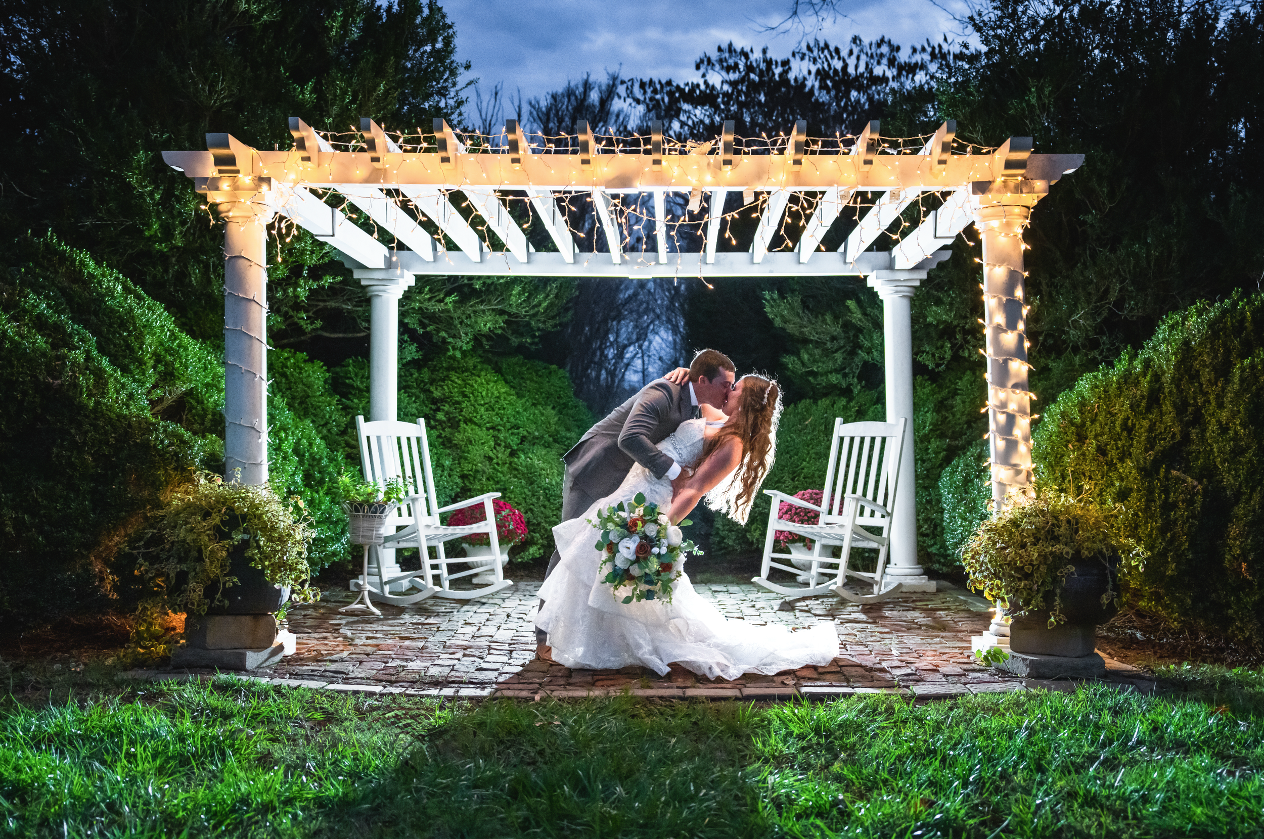 A newlywed couple sharing a kiss under a lit pergola at night. The bride is holding a bouquet, and the groom is dipping her in a romantic pose. The scene is illuminated by string lights, with greenery and two white rocking chairs on either side.