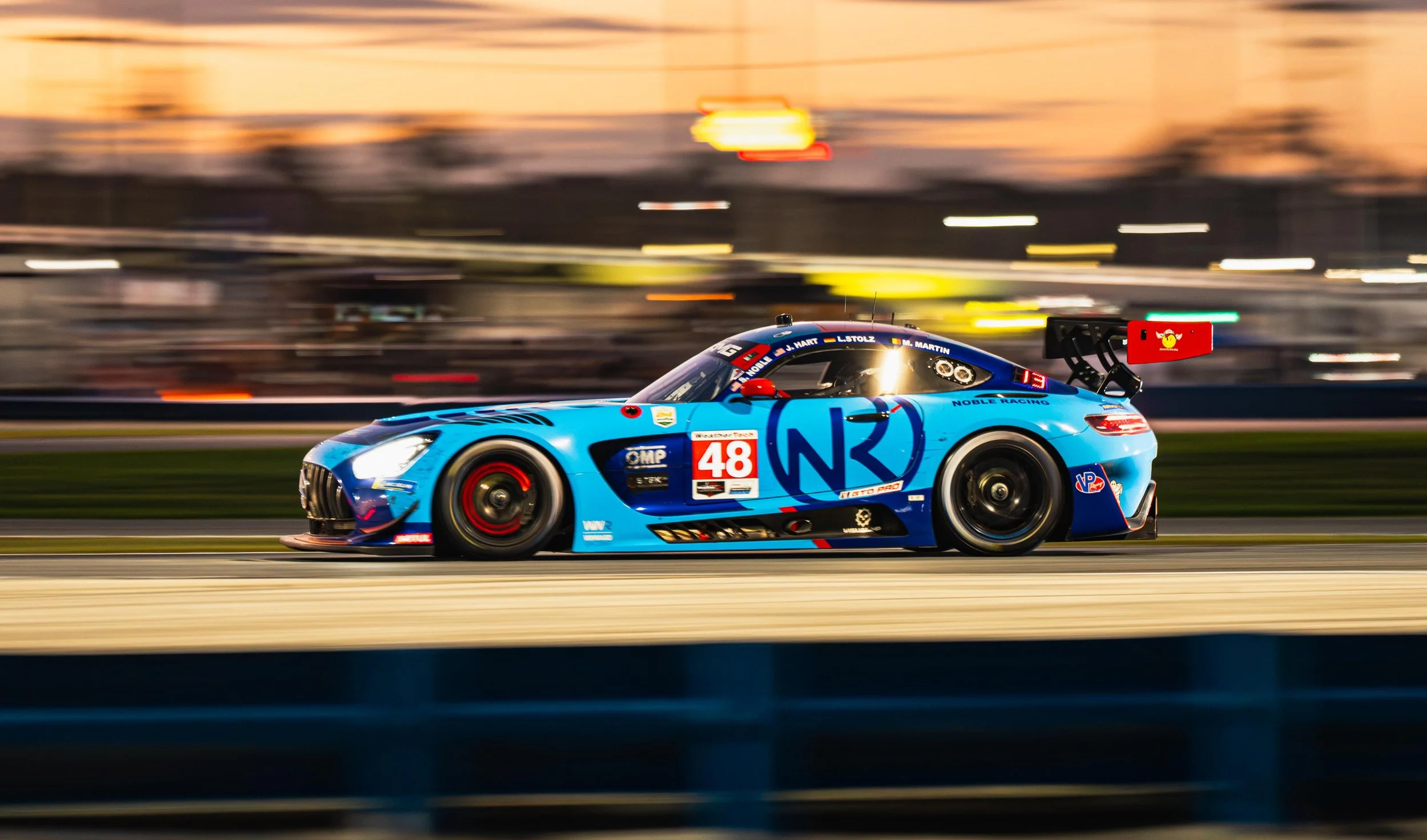 Blue race car with number 48 on the side is racing on a track during sunset, with a blurred background indicating high speed.