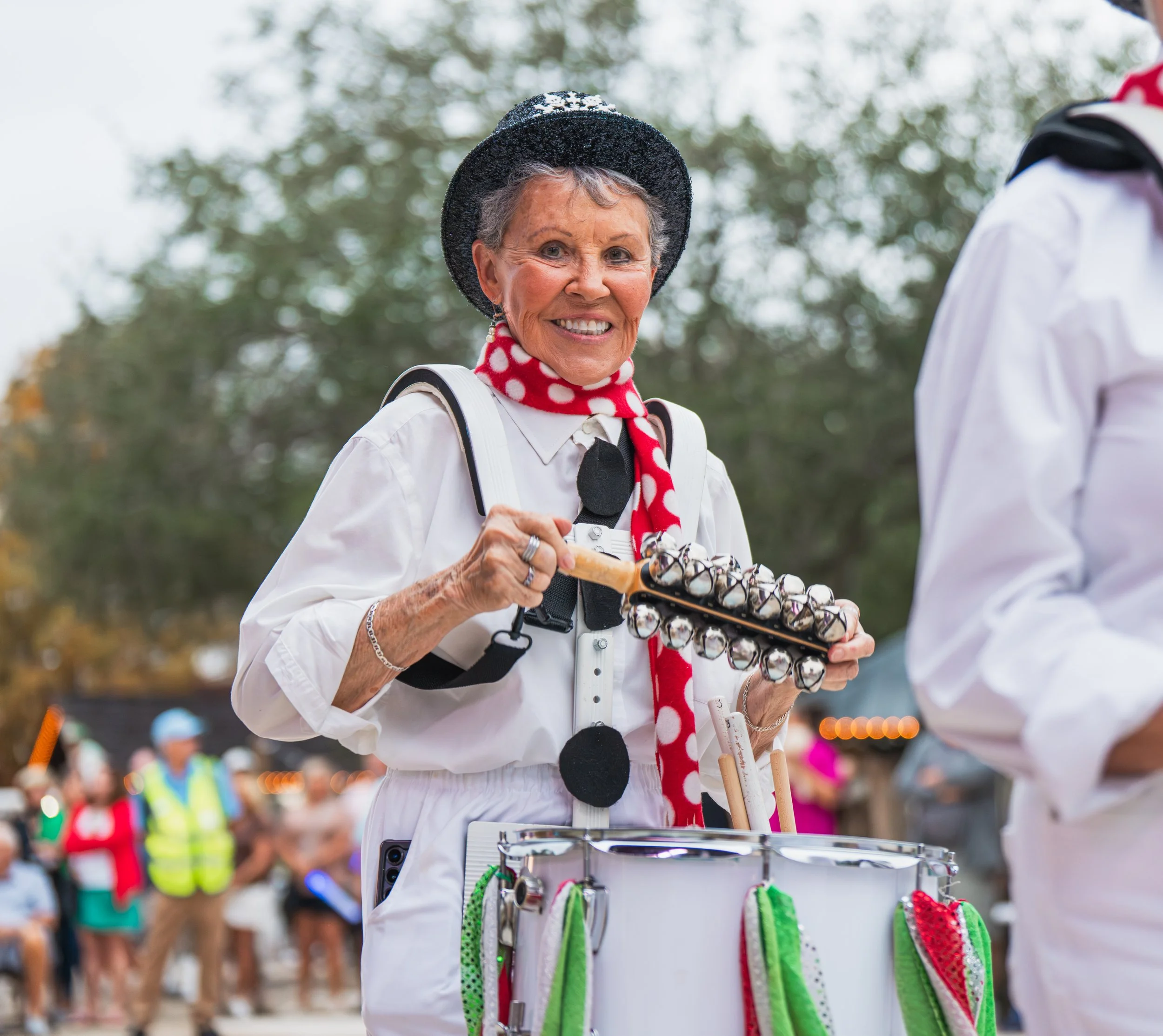 A smiling woman in a white outfit with a red polka dot scarf and black hat playing bells at an outdoor event, surrounded by other people.