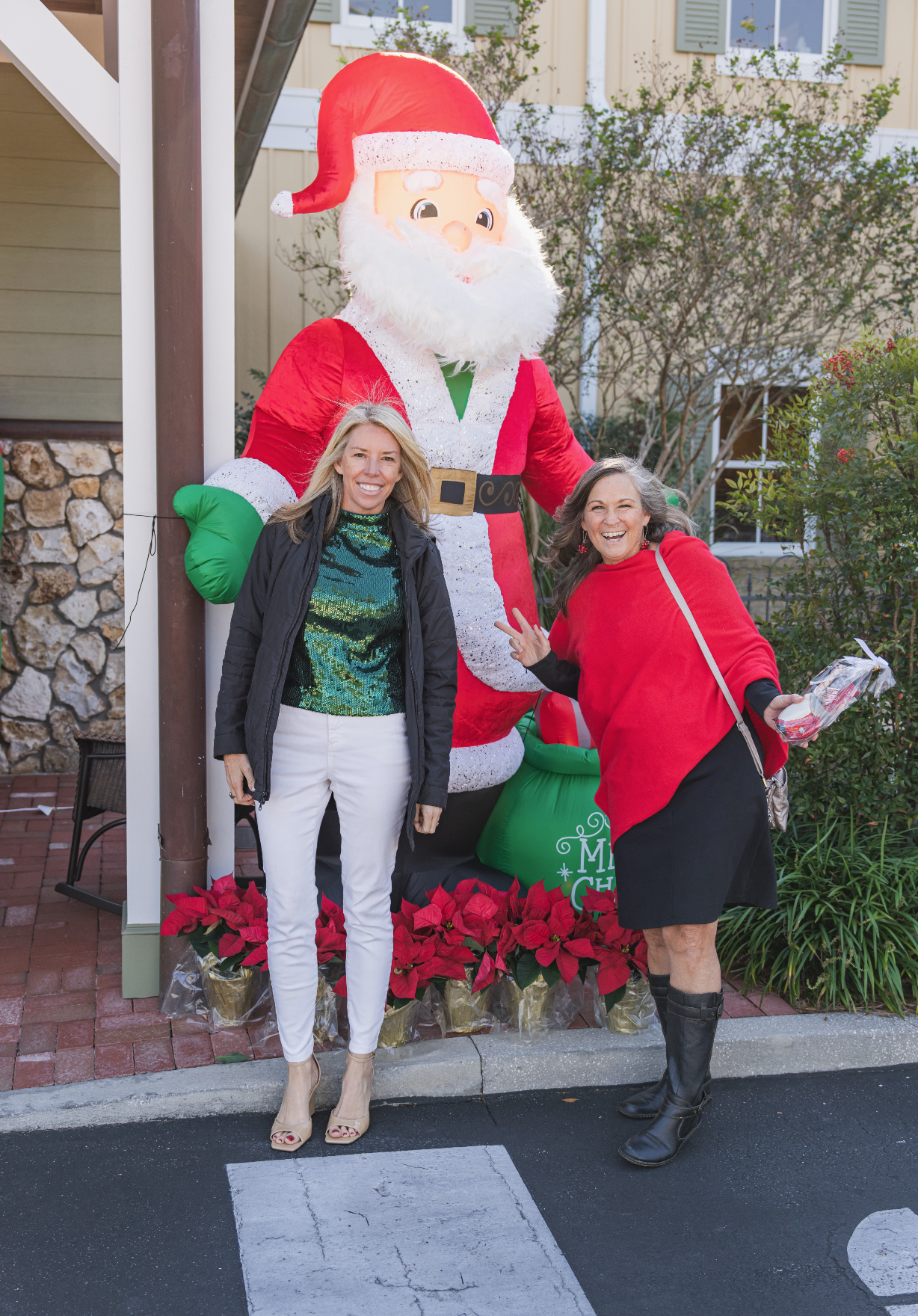 Two women smiling and posing in front of a large inflatable Santa Claus decoration. One woman is wearing a black jacket and white pants, and the other is wearing a red sweater and black skirt. They are standing on a sidewalk with festive poinsettia p