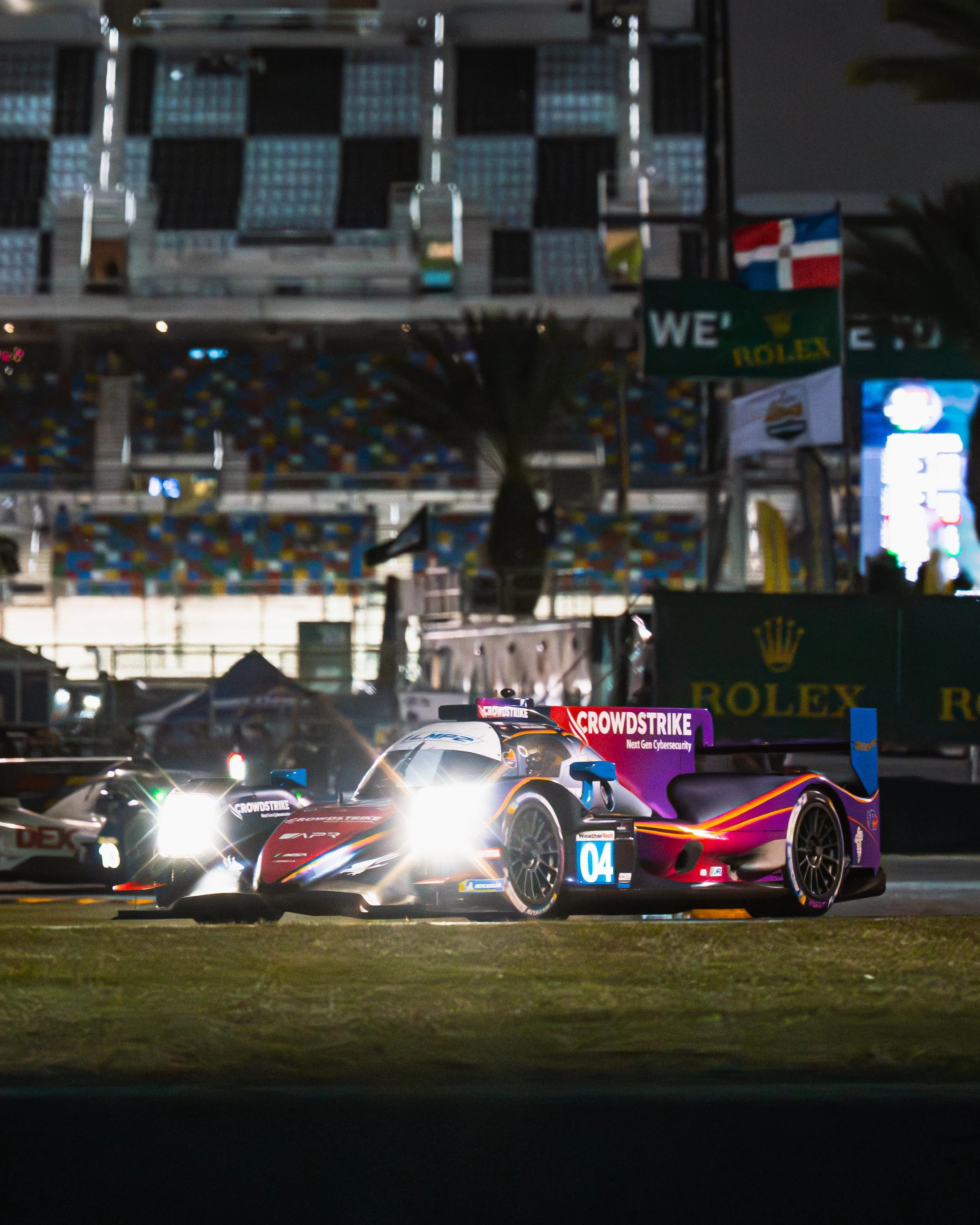 A race car with bright headlights at night, on a race track, with a blurry background of grandstands and advertisements.