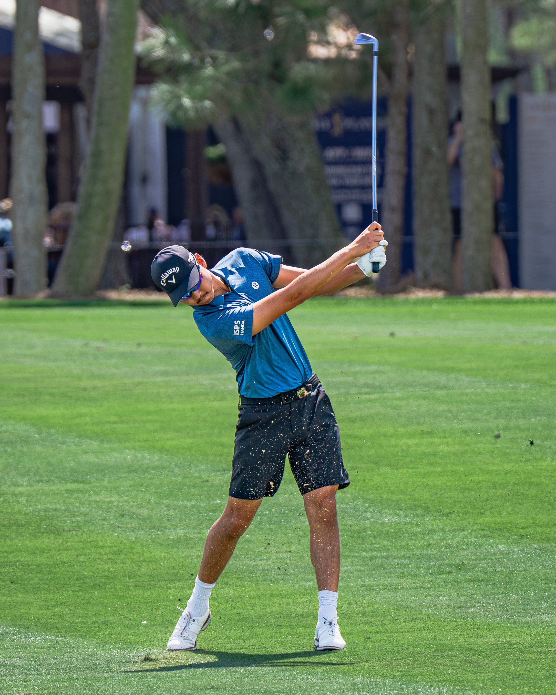 A male golfer is swinging a golf club on a golf course with trees in the background.