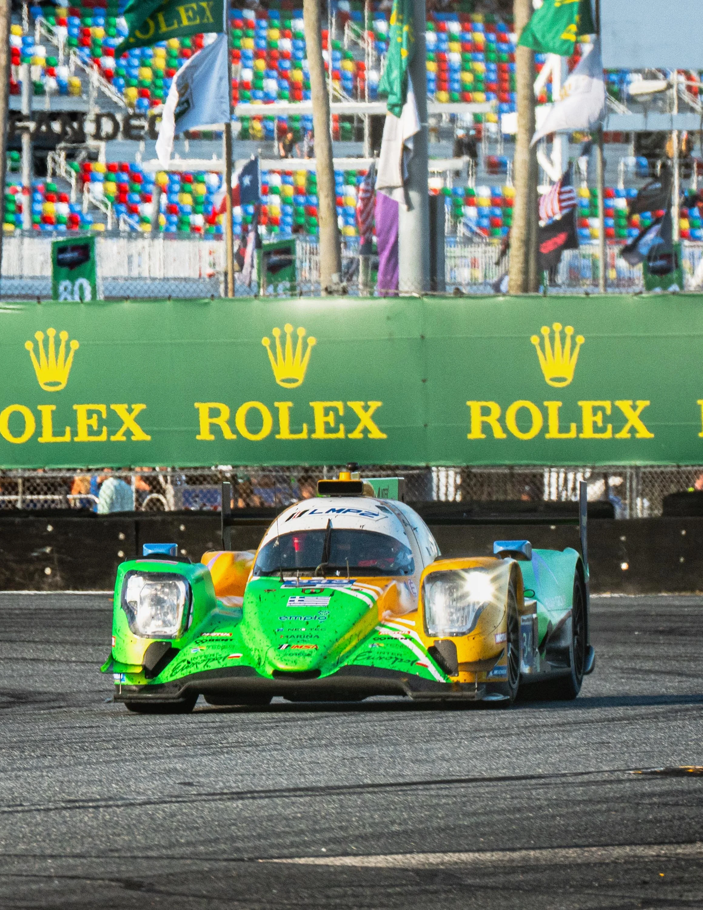 A race car with green and yellow livery speeding on a track, with a green Rolex advertisement banner and colorful spectators' stands in the background.