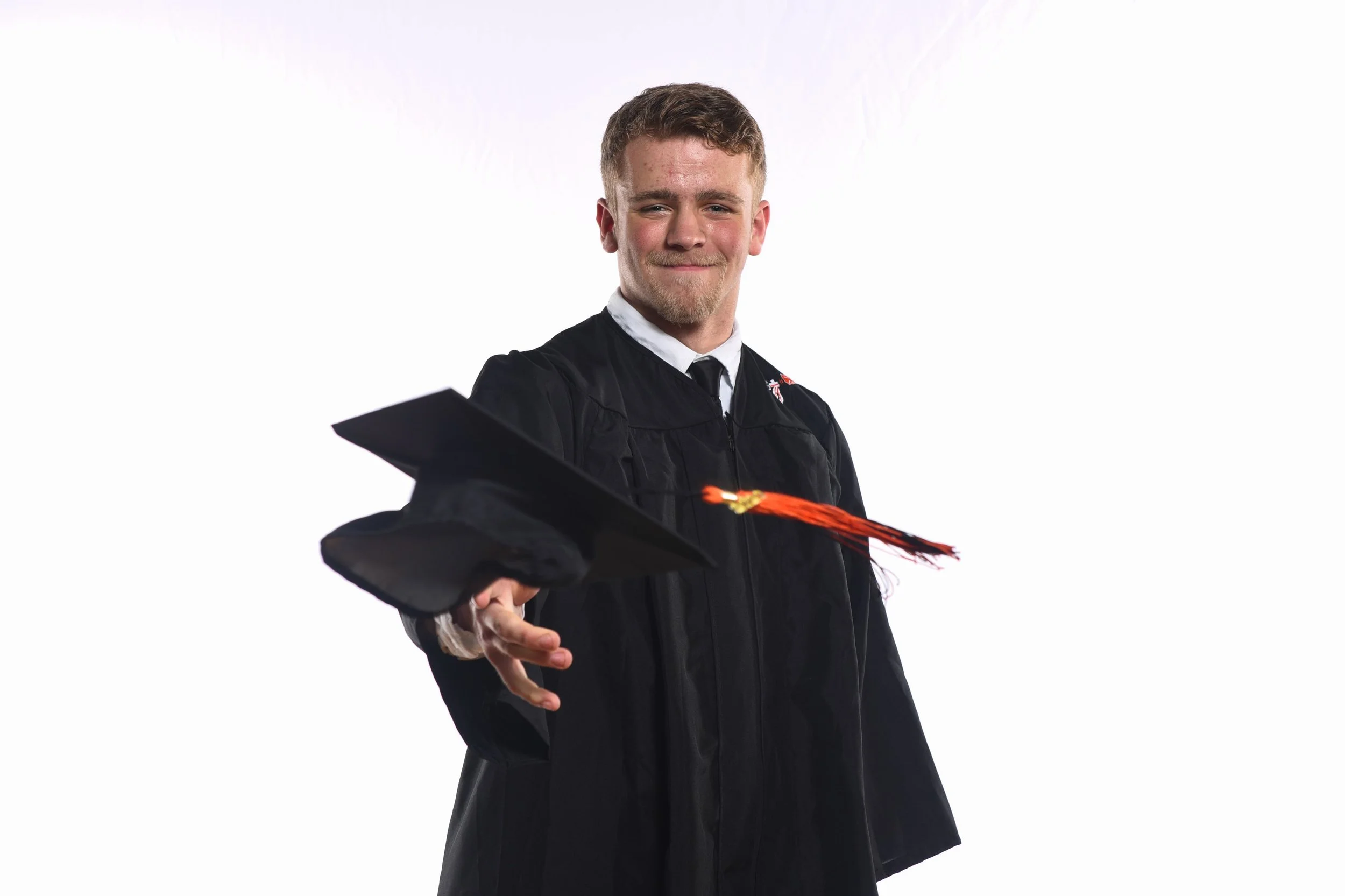 Graduation photography. Senior photography central Florida. A young man in a graduation cap and gown throws his cap in the air, smiling, against a plain white background.