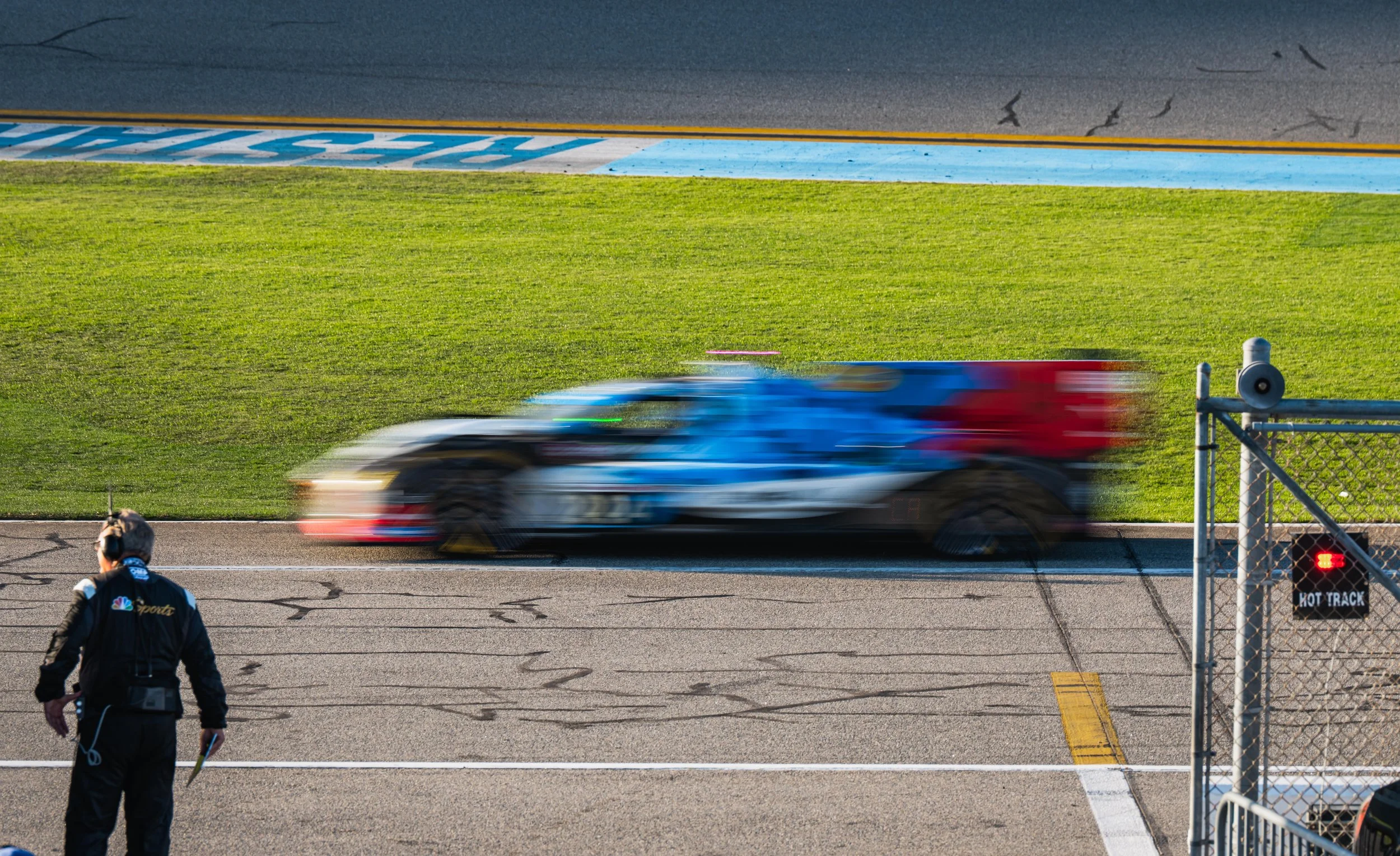 A blurred race car passing the pit lane of a race track with a crew member standing in the foreground, wearing a headset and holding a notepad.