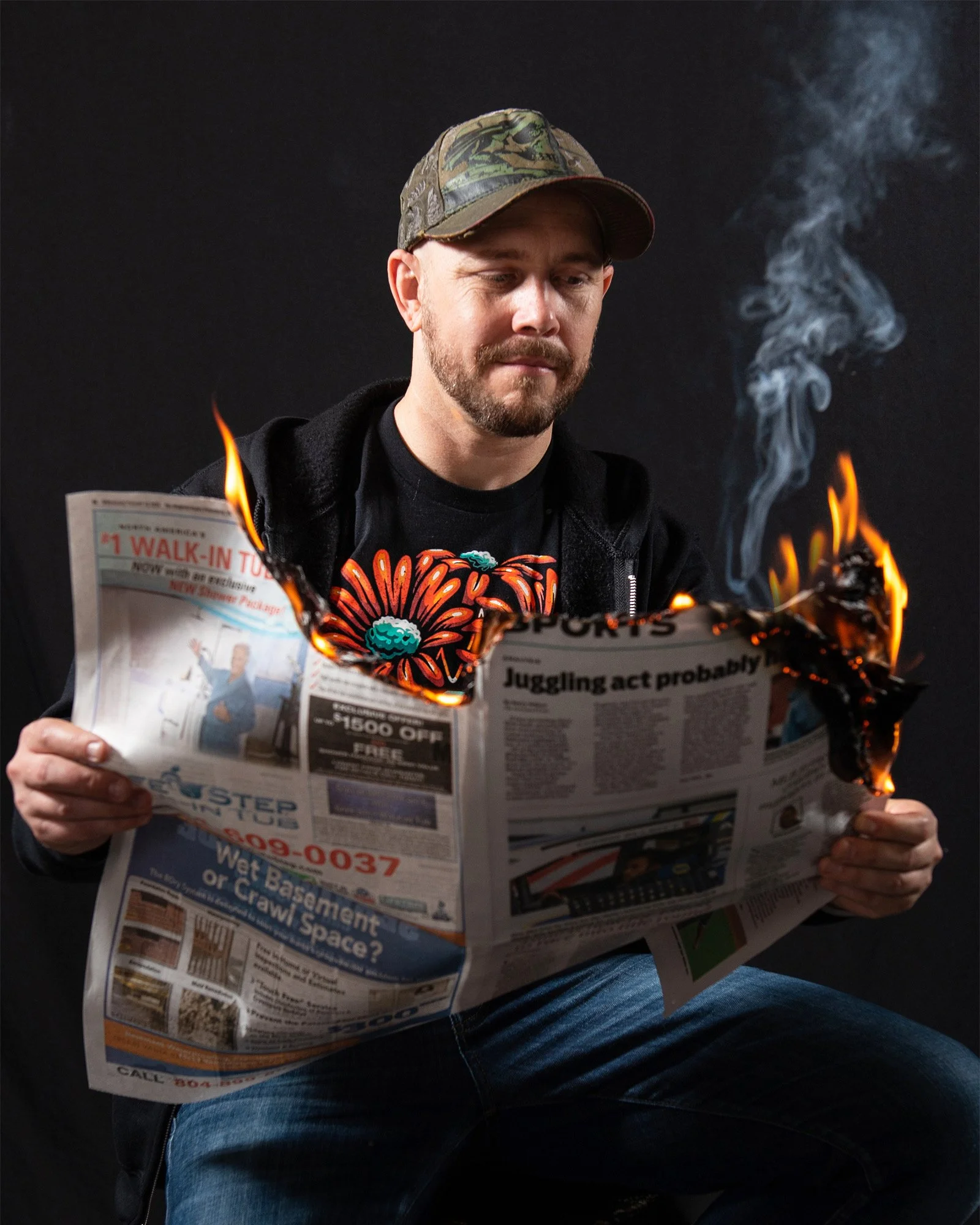 Jesse Smith Tattoo Artist wearing a cap and black hoodie, sitting and holding a burning newspaper with visible flames and smoke against a dark background.