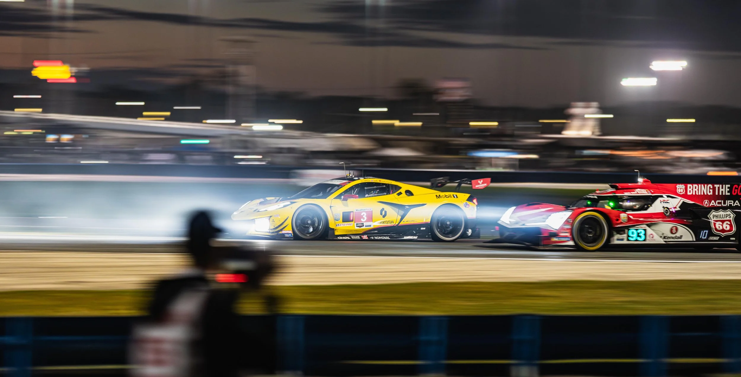 Nighttime race featuring two racing cars on a track, one yellow with black accents and one red, white, and black, with motion blur indicating high speed.