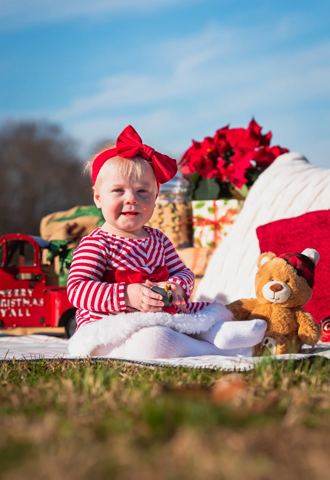 A young girl in Christmas attire, sitting outdoors on a blanket with a teddy bear, Christmas decorations, and gift boxes around her.