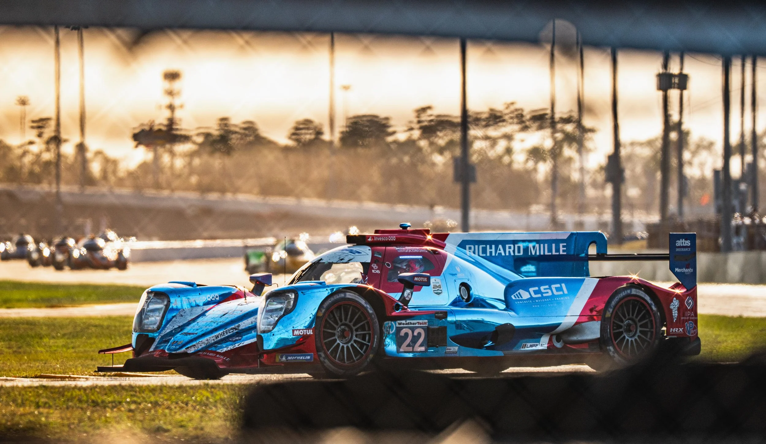 A racing car with blue, red, and white livery on a racetrack, with the driver's helmet visible through the windshield, and multiple cars blurred in the background during sunset.