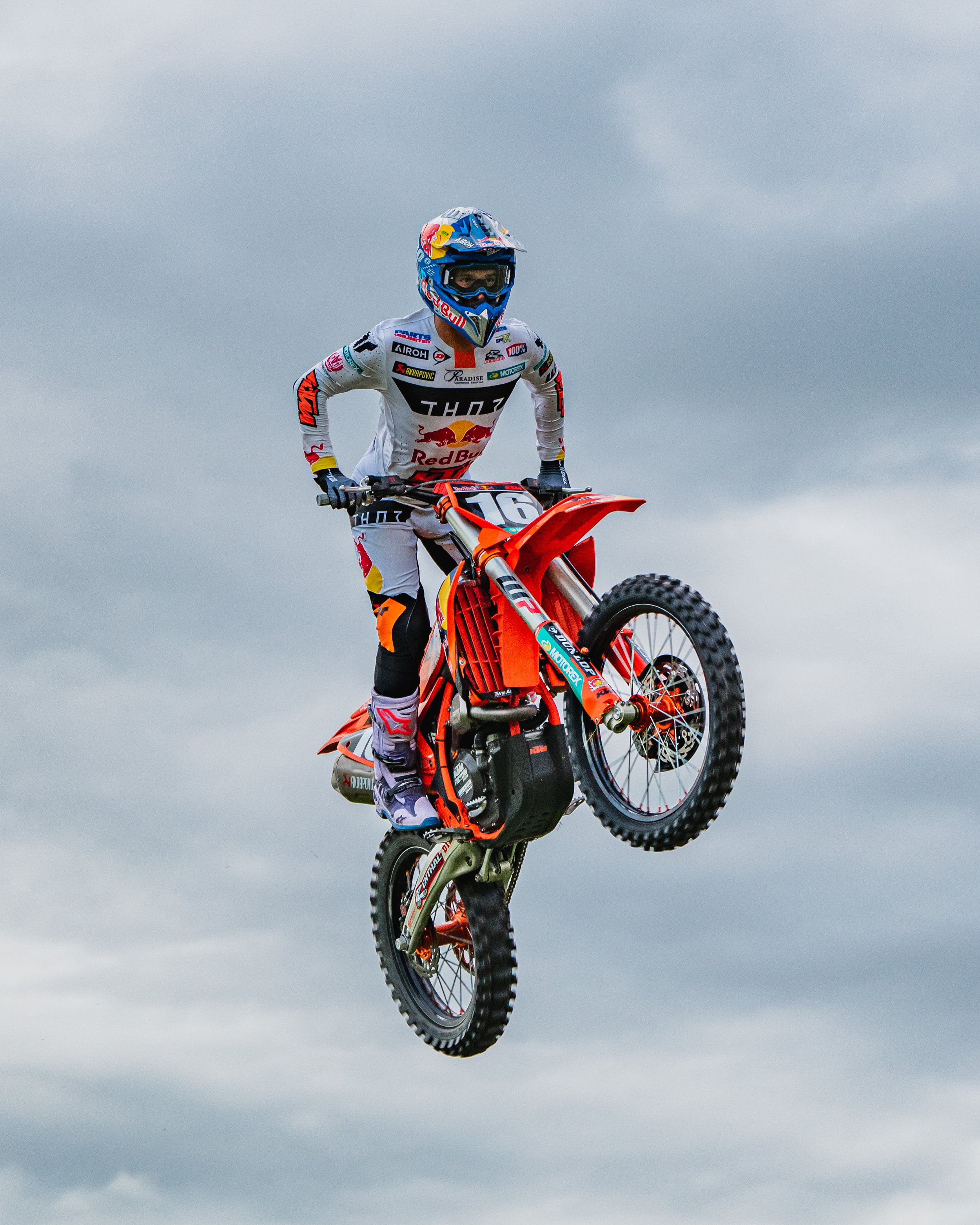Motocross rider in full gear mid-air on an orange dirt bike against a cloudy sky.