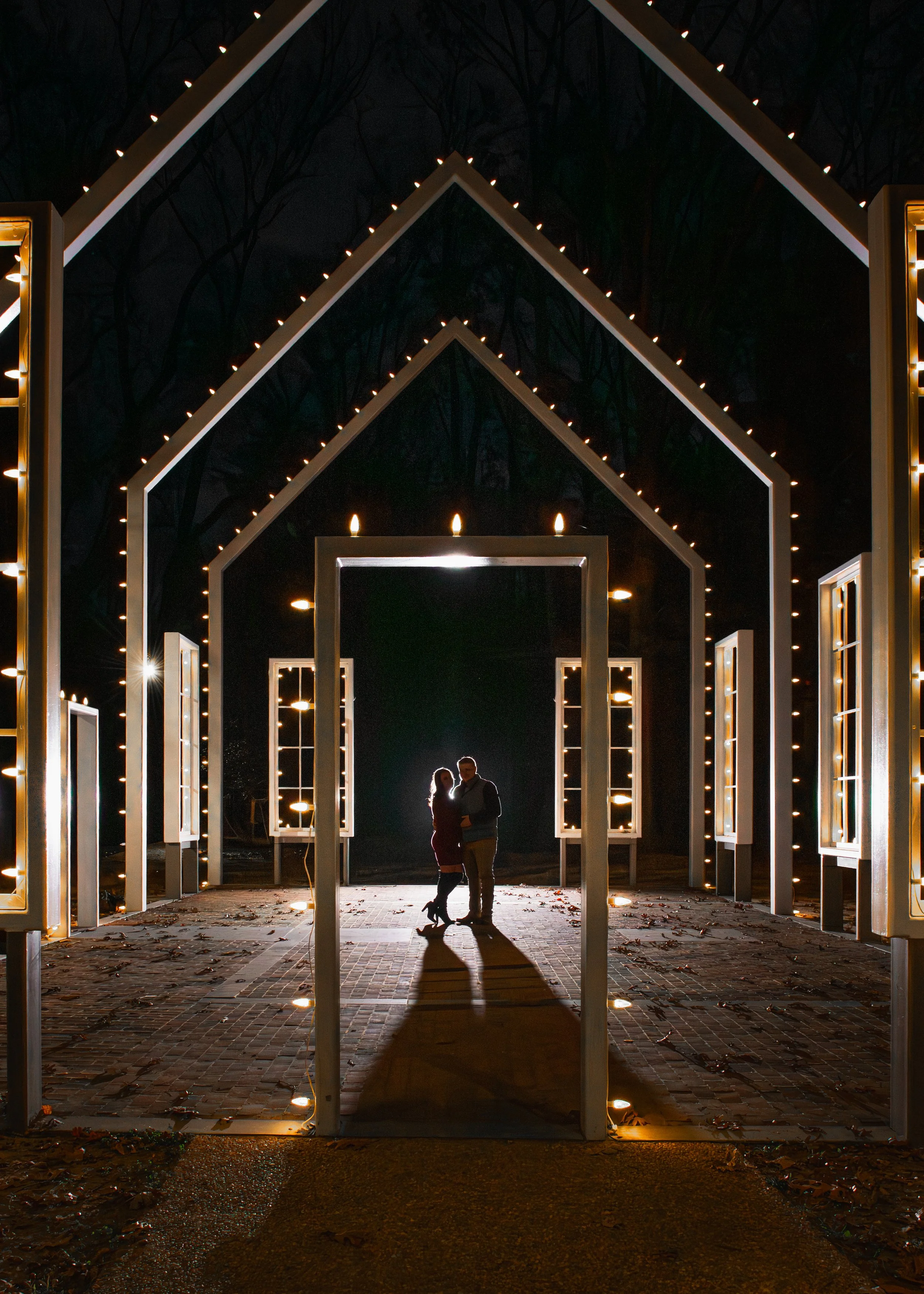 Silhouettes of a couple standing close together in front of illuminated archways at night, with decorative lighting and a dark background with faint trees. Polegreen Church, Virginia