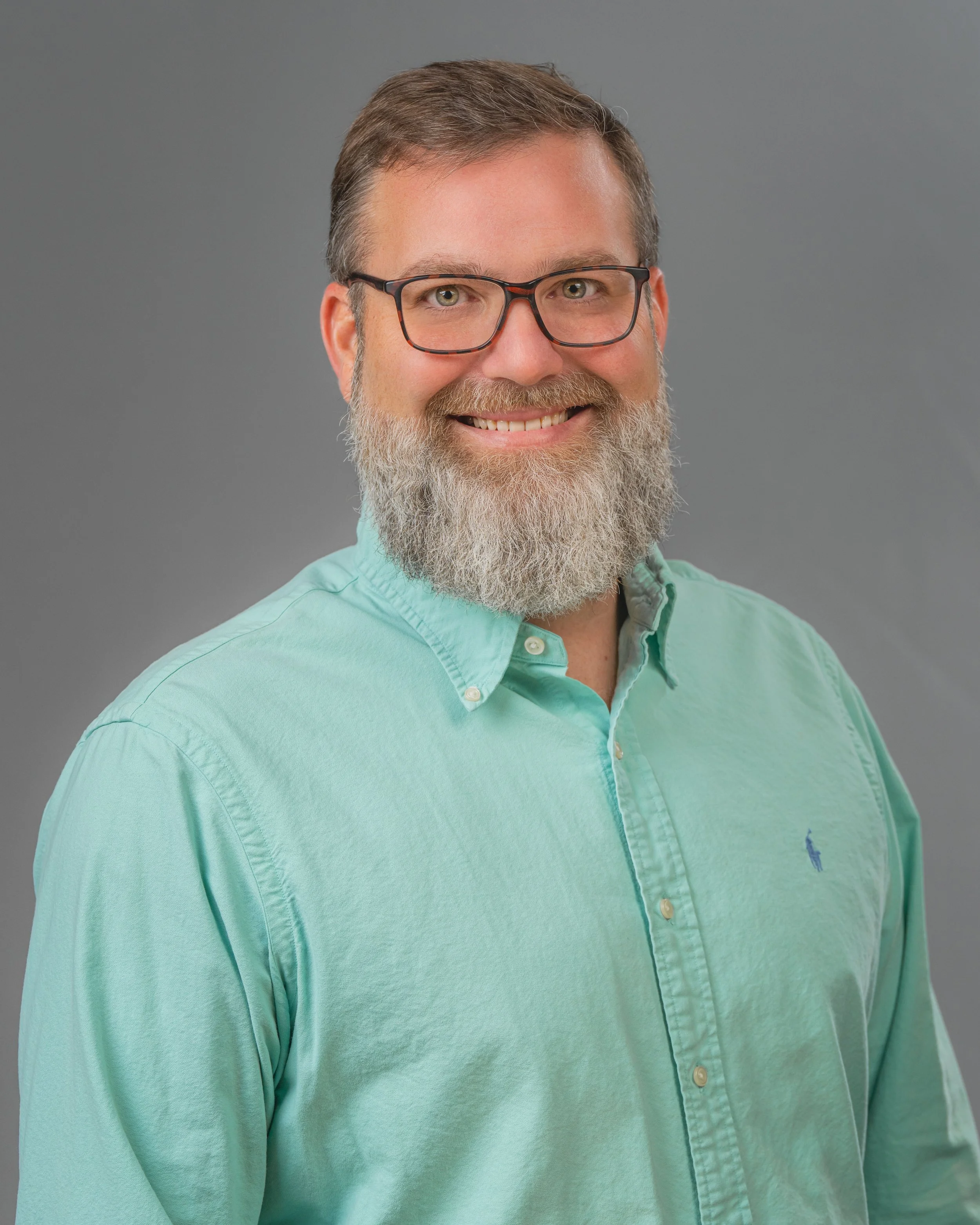 Portrait photography, Central Florida. Corporate headshots. Portrait of a man with glasses, a beard, smiling, wearing a mint green button-up shirt, against a gray background.