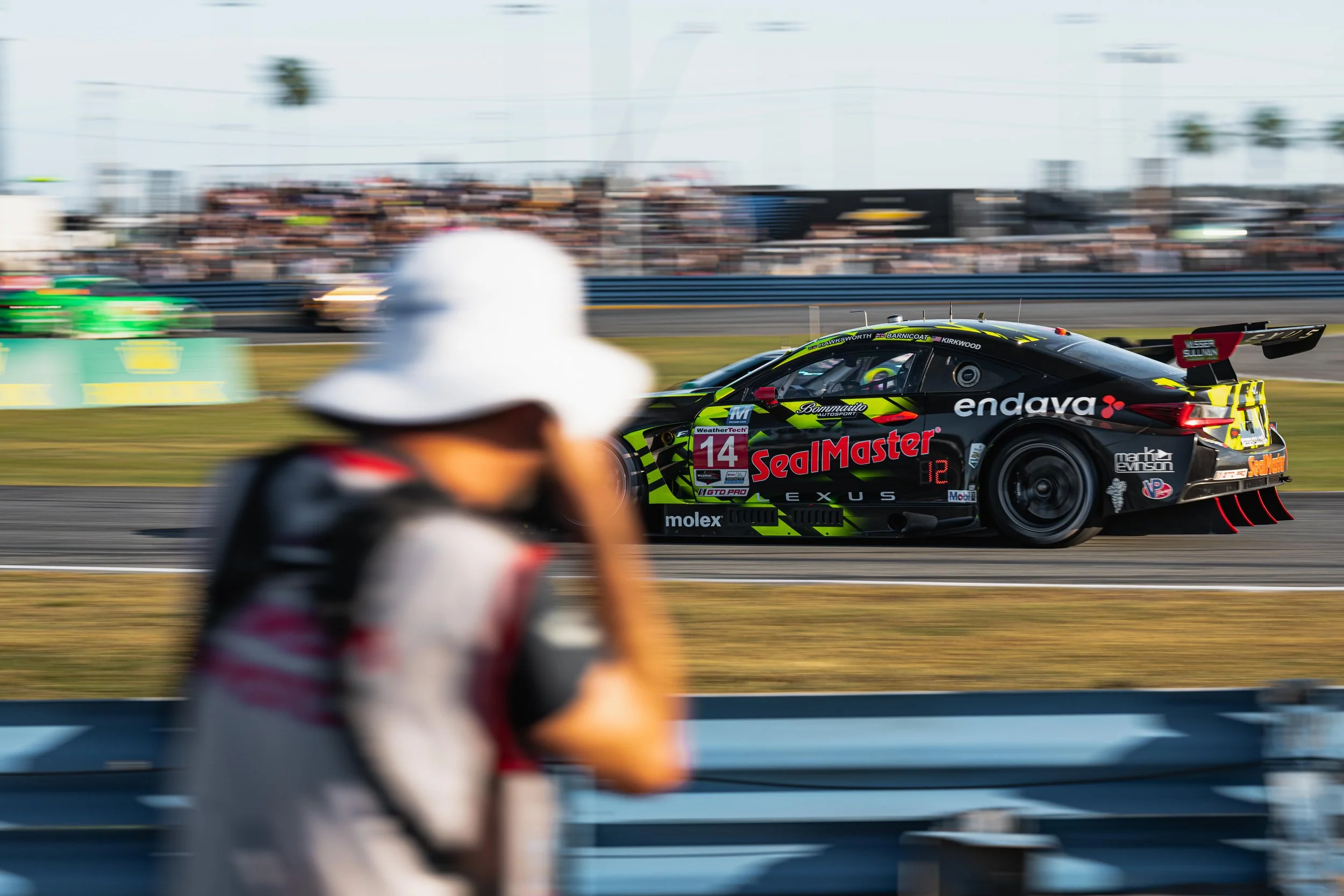 Blurred person wearing a white hat and black camera strap in the foreground, blurred background of racing cars on a track, with a black and neon green race car in sharp focus.
