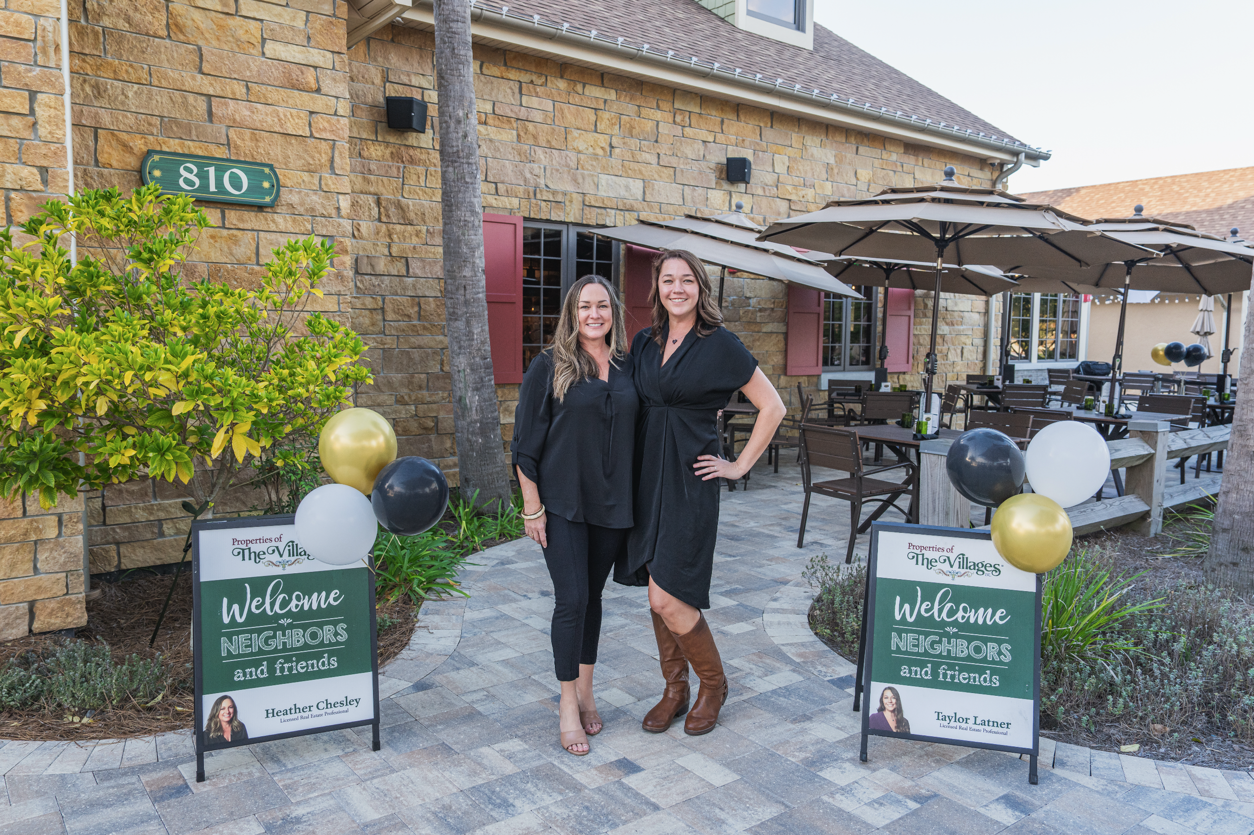 Two women standing outside a building with a sign that reads 'Welcome NEIGHBORS and friends' in front of a patio with tables and umbrellas. One woman is wearing a black dress and brown boots, and the other is wearing a black blouse and pants.