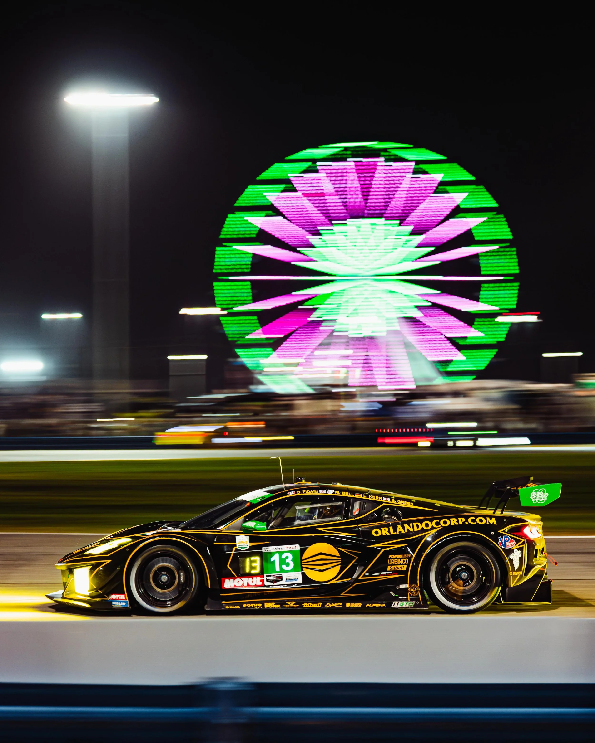 A race car speeding on a track at night, with a brightly lit Ferris wheel in the background illuminated with purple and green lights.