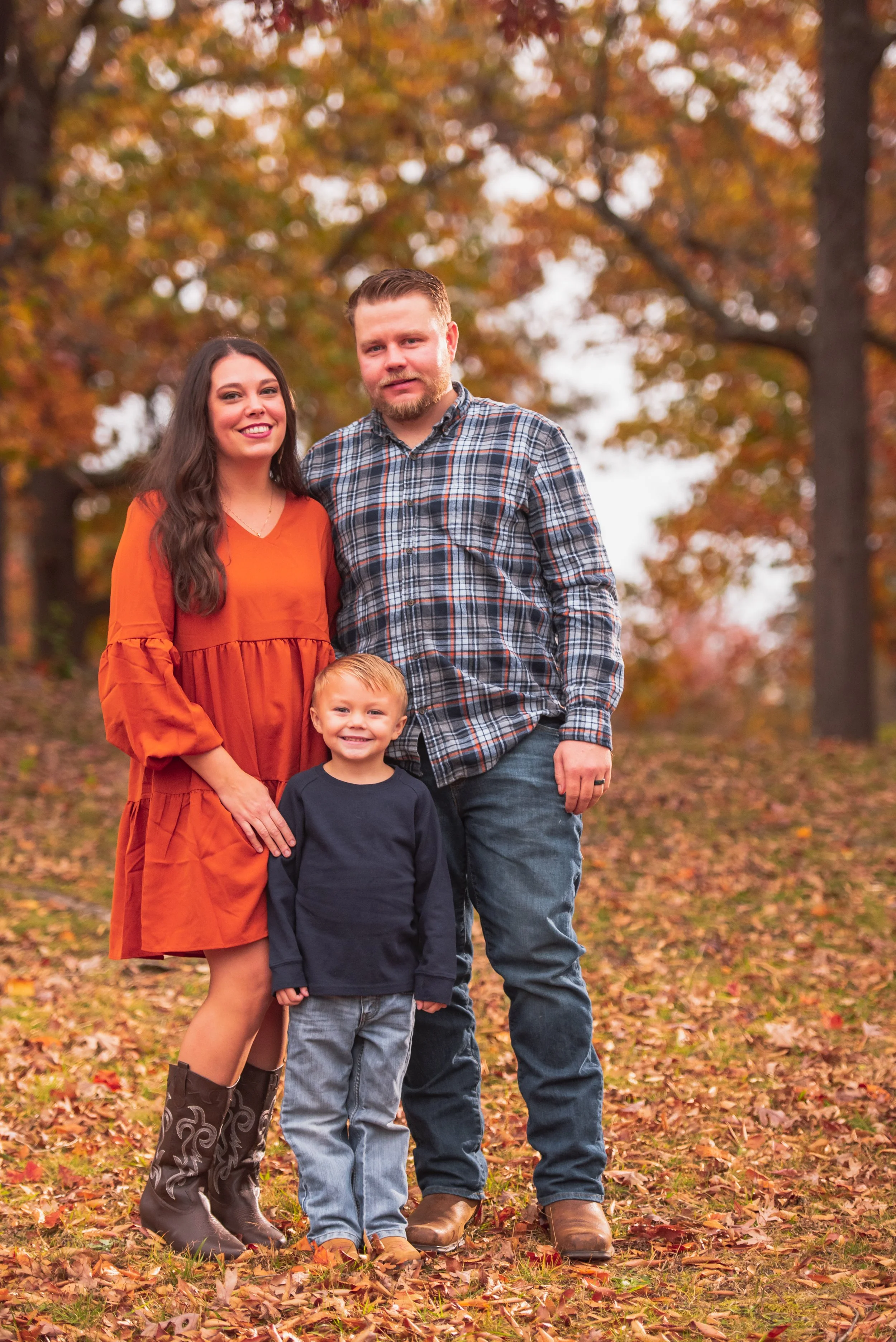 A family of three standing outdoors during fall, with colorful autumn trees and fallen leaves. The woman is wearing an orange dress with black boots, the man is in a plaid shirt and jeans, and the young boy is dressed in a navy blue sweatshirt and je
