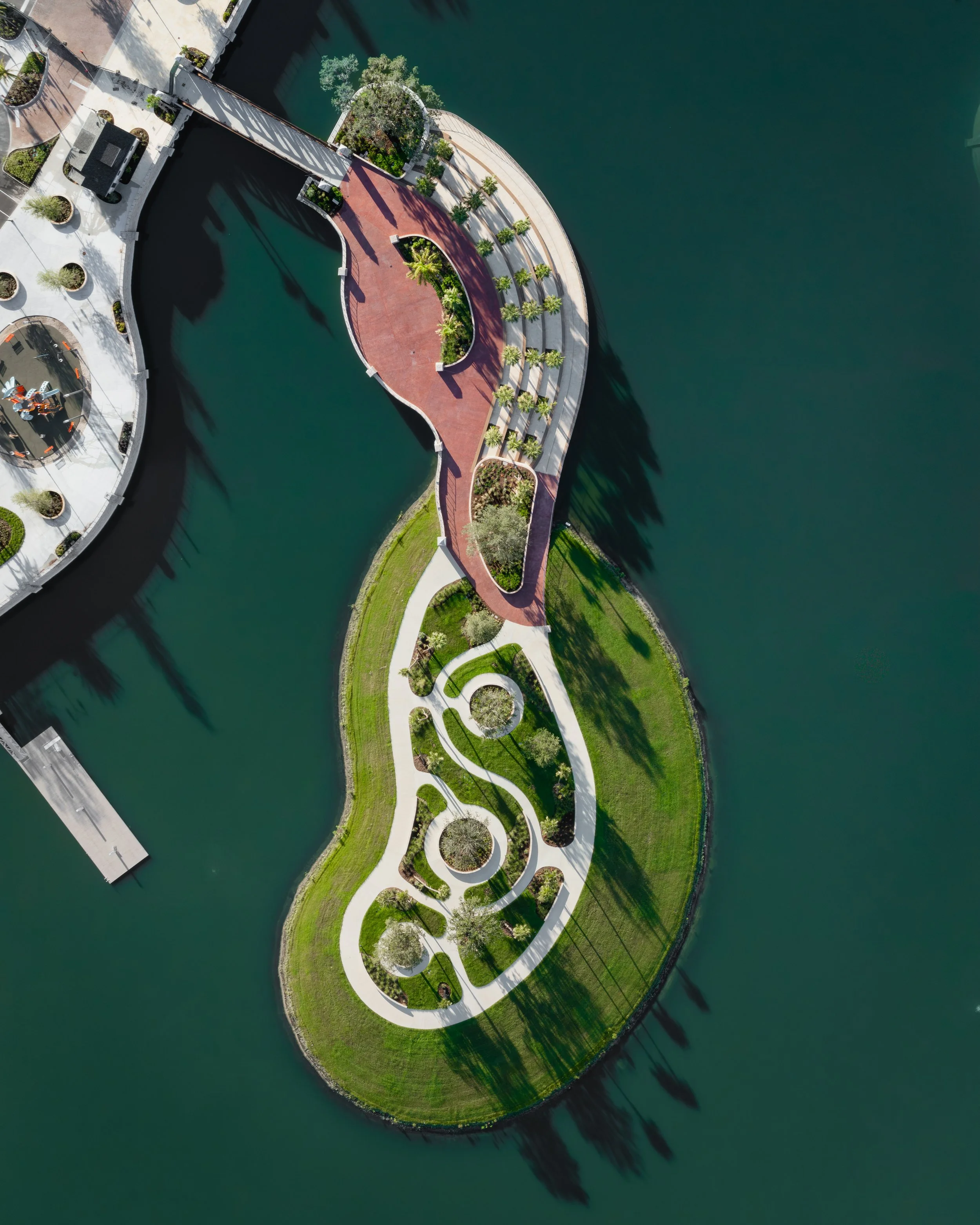 Aerial view of a park surrounded by water, featuring walking paths, greenery, and landscaped areas with trees and shrubs.