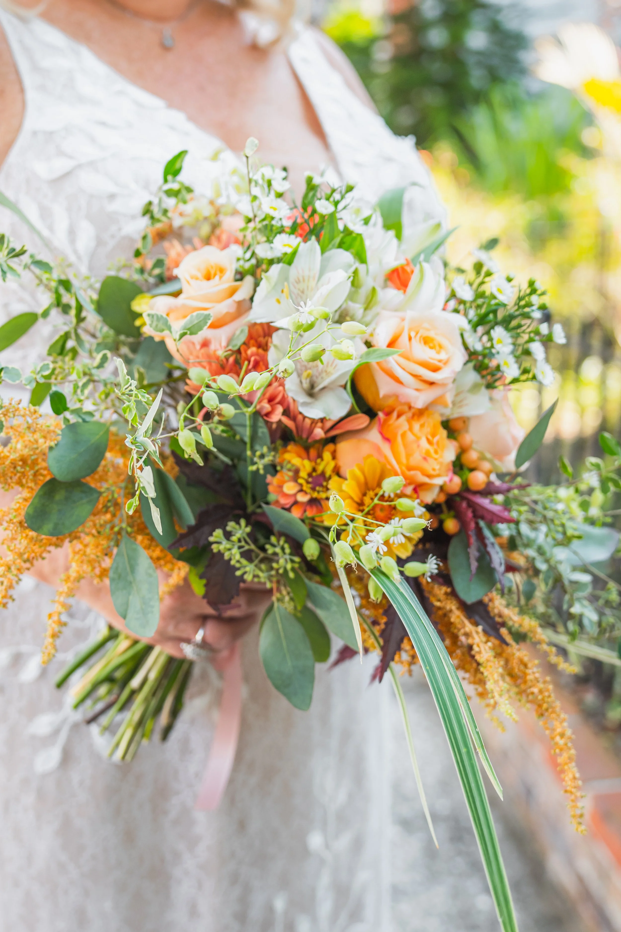 A woman in a lacy white dress holding a large colorful bouquet of flowers outdoors.