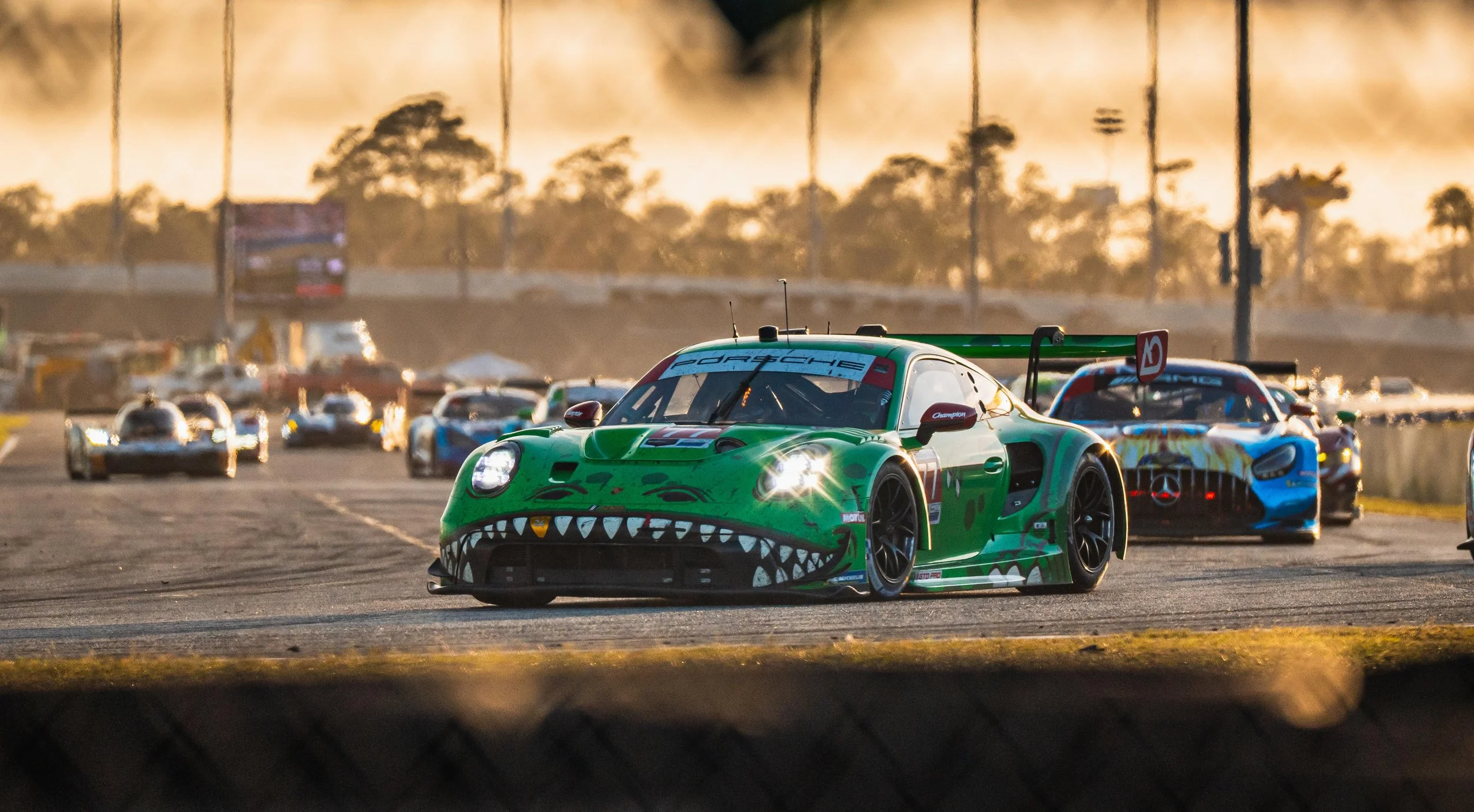 Race cars on a track at sunset, with the leading car featuring a green paint job with a shark mouth design.