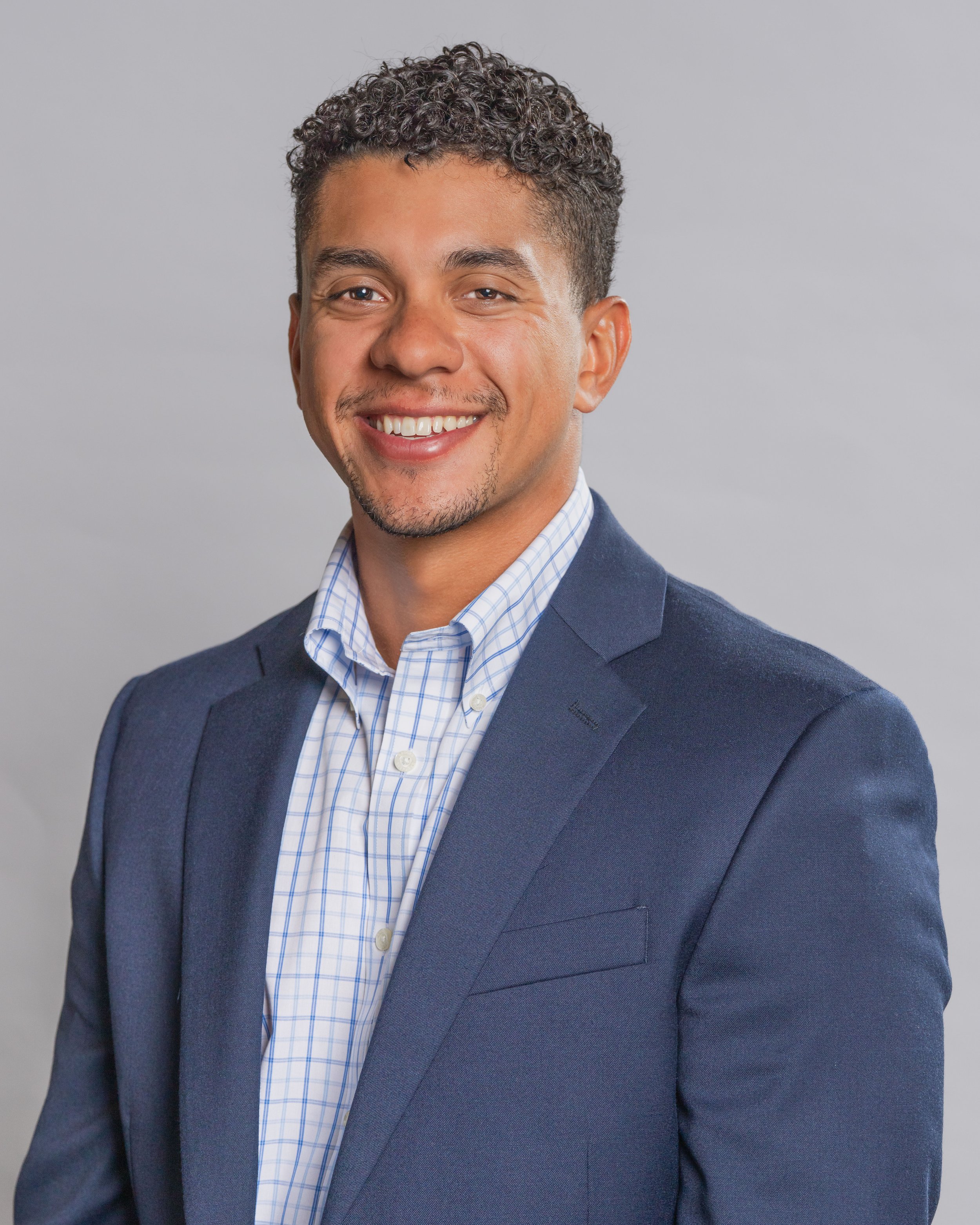 Portrait photography, Central Florida. Corporate headshots. A smiling man with short, curly dark hair, wearing a blue blazer and a blue checkered button-down shirt, against a plain gray background.