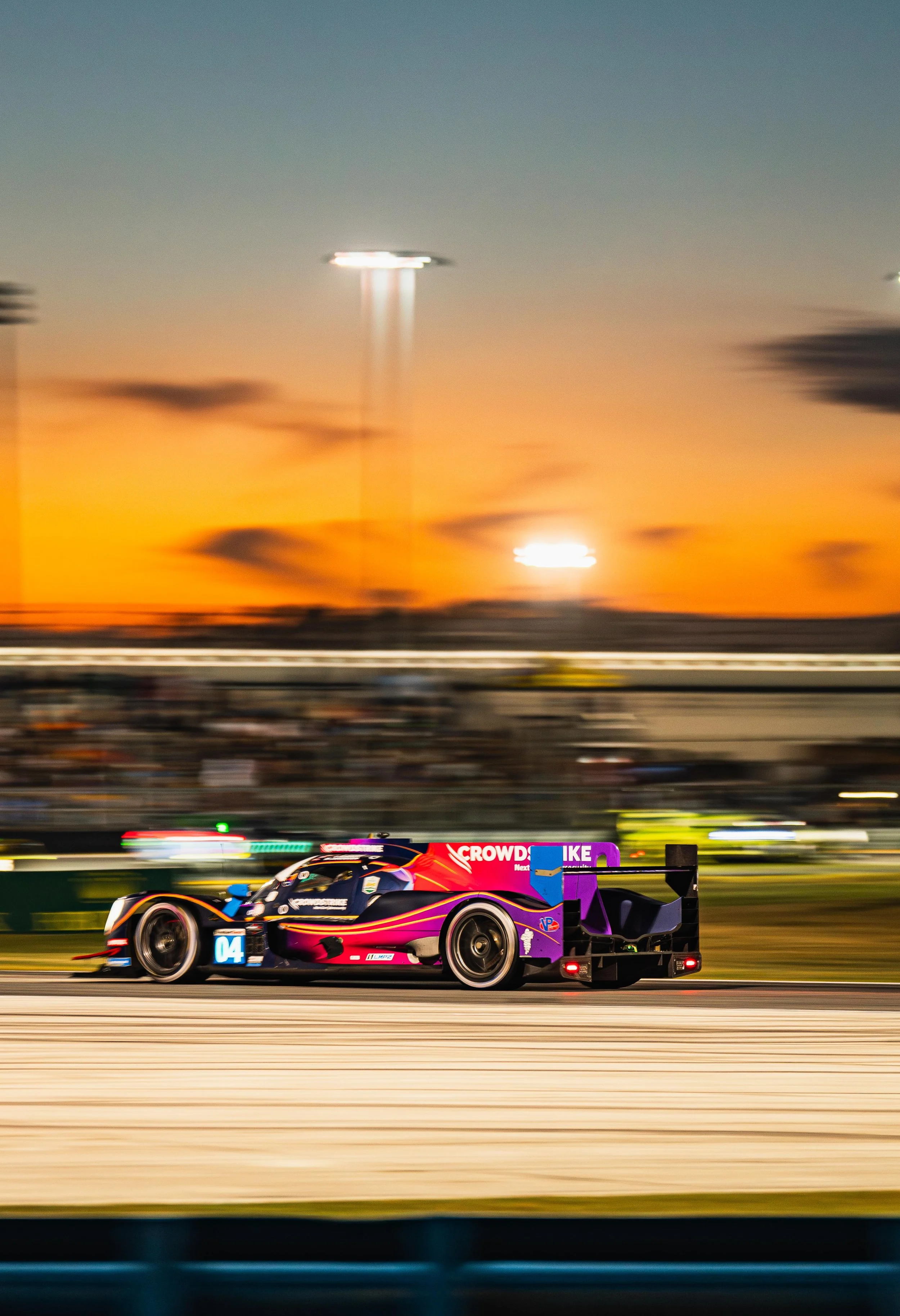 A race car speeding on a track during sunset, with the sky displaying orange and blue hues and stadium lights in the background.