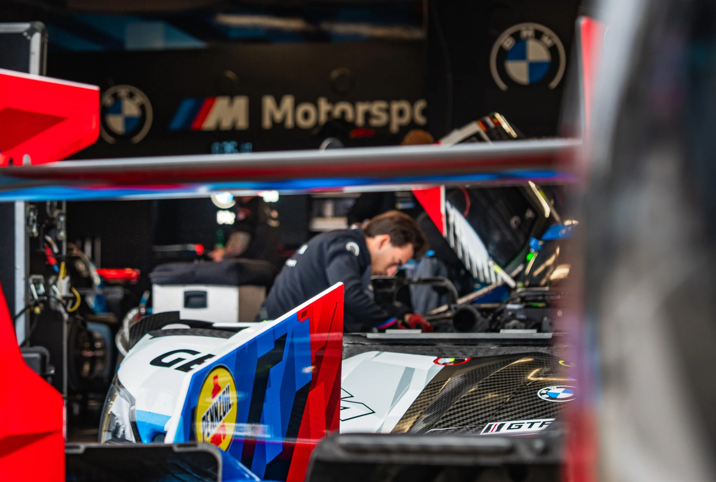 Motorsport garage with a mechanic working on a race car, surrounded by various tools and equipment. The garage features BMW and other racing logos.