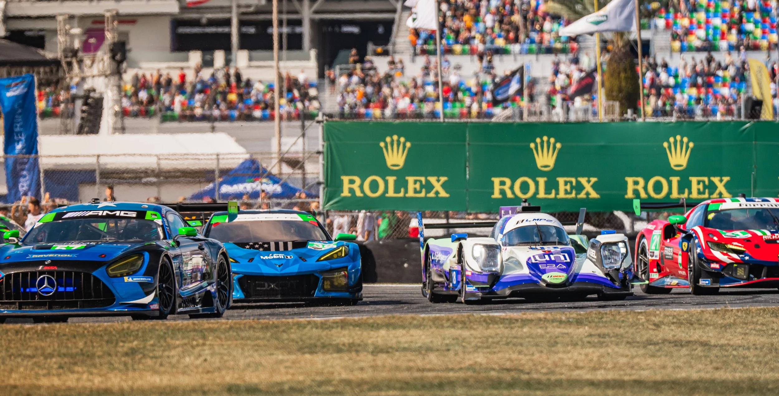 Four race cars on a track with a crowded grandstand and green Rolex banners in the background.