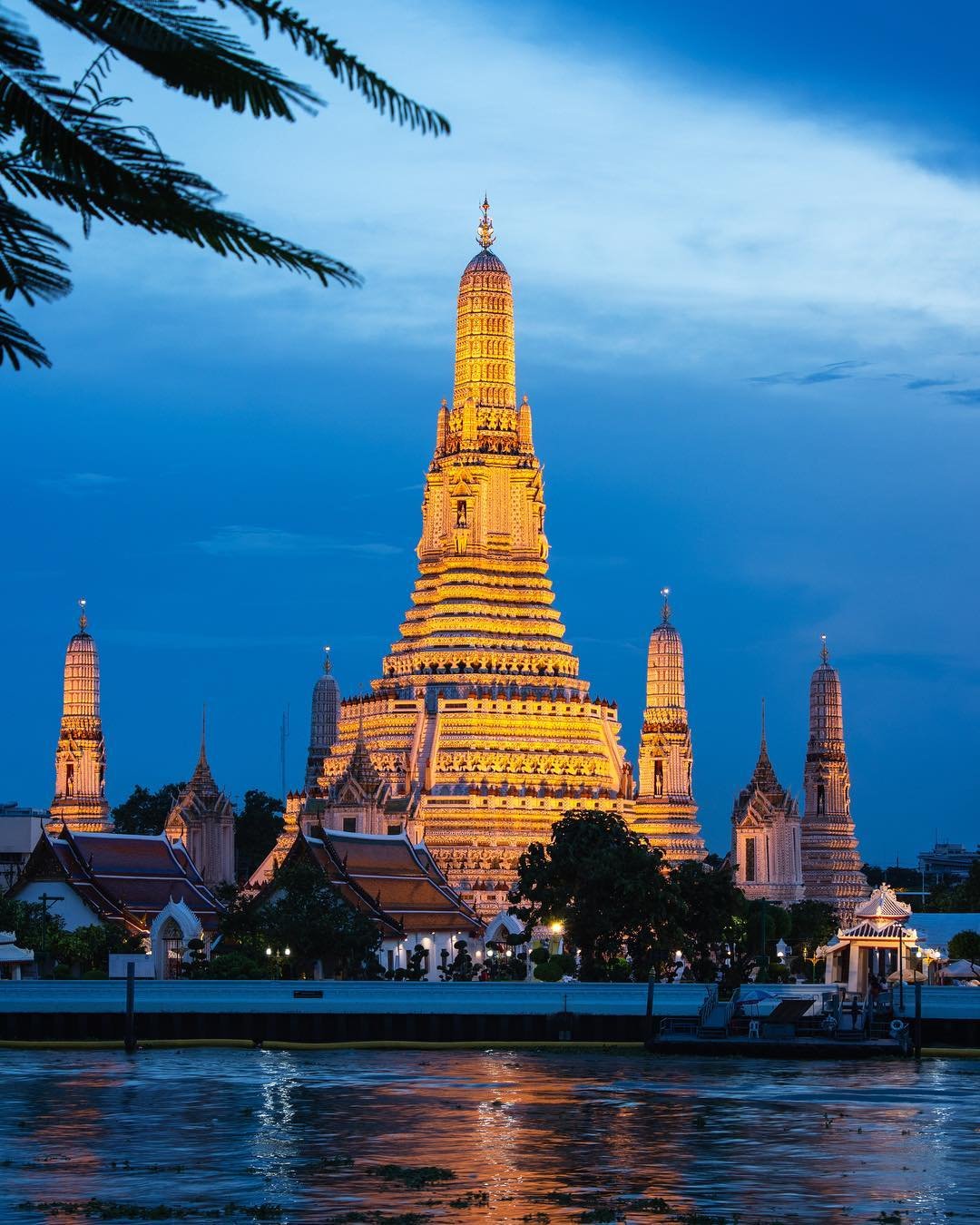 Wat Arun temple illuminated at dusk with a reflection on the water, trees, and a partly cloudy sky in the background.