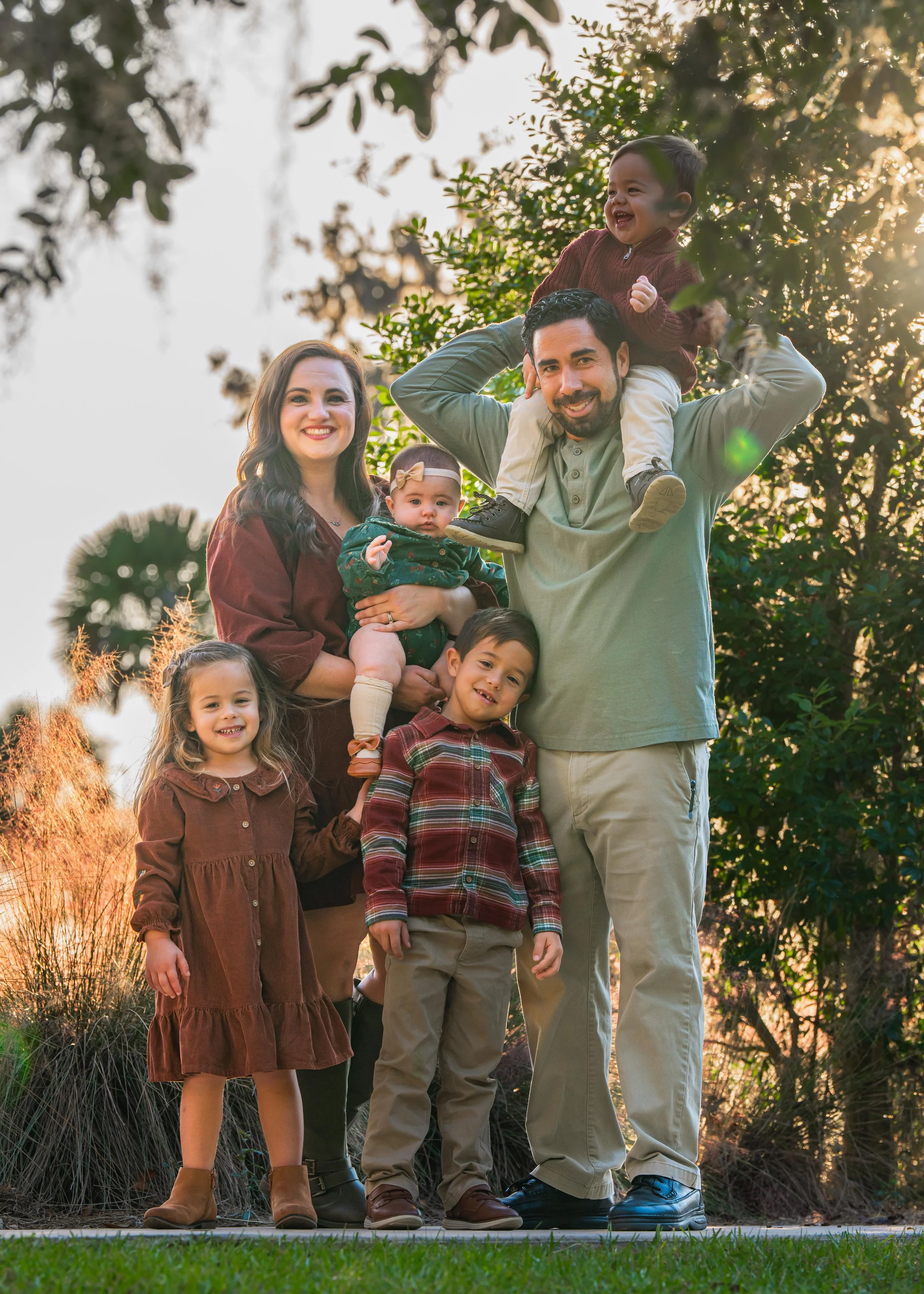 A family of seven posing outdoors during sunset, with trees in the background. The father is carrying a young boy on his shoulders, the mother is holding a baby girl, and two other children stand in front, smiling.