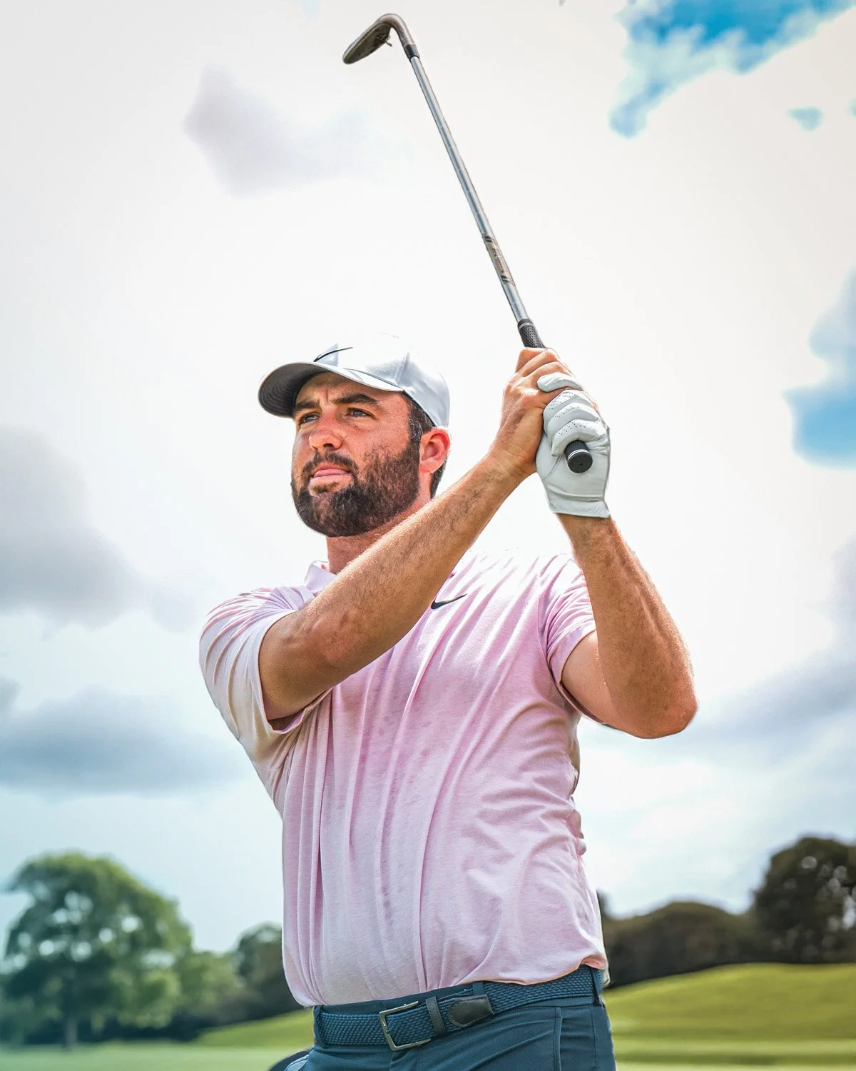 Scottie Scheffler swinging a golf club on a golf course with trees and a cloudy sky in the background. 