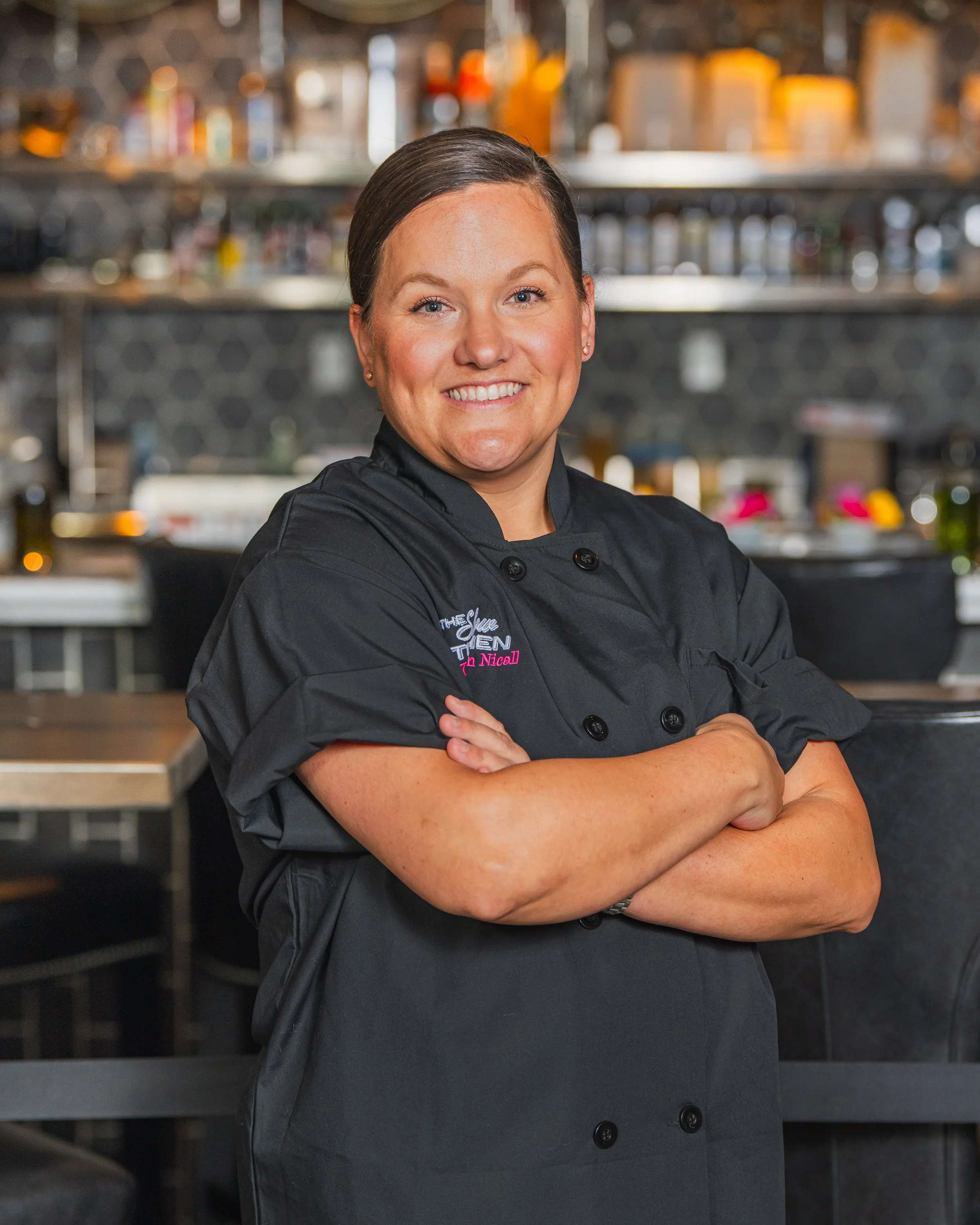 Smiling female chef in black uniform with arms crossed in a restaurant kitchen.
