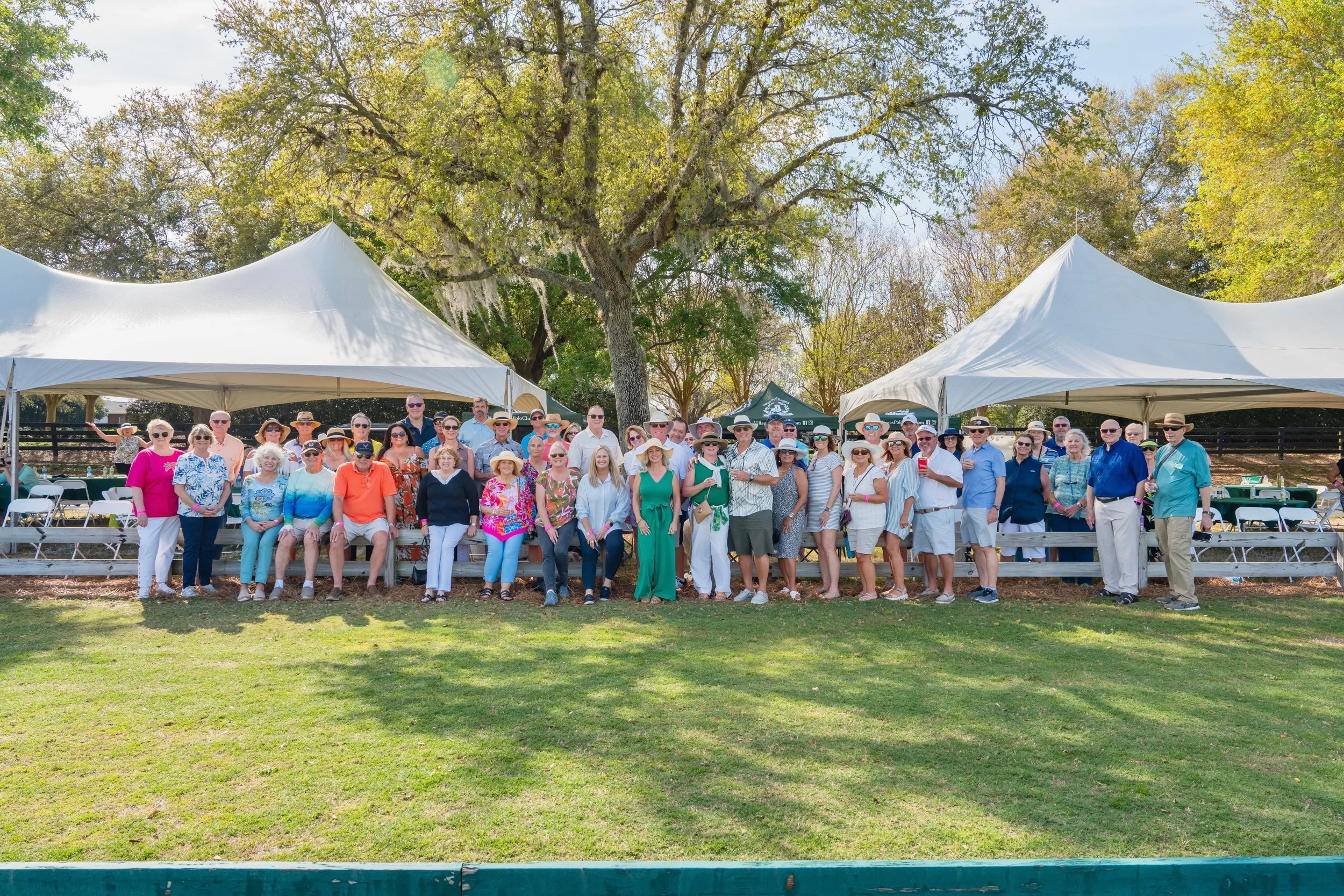 A large group of people posing for a photo outdoors on a sunny day, with white tents and trees in the background. The group includes men and women dressed in casual and summer clothing, some wearing hats and sunglasses.