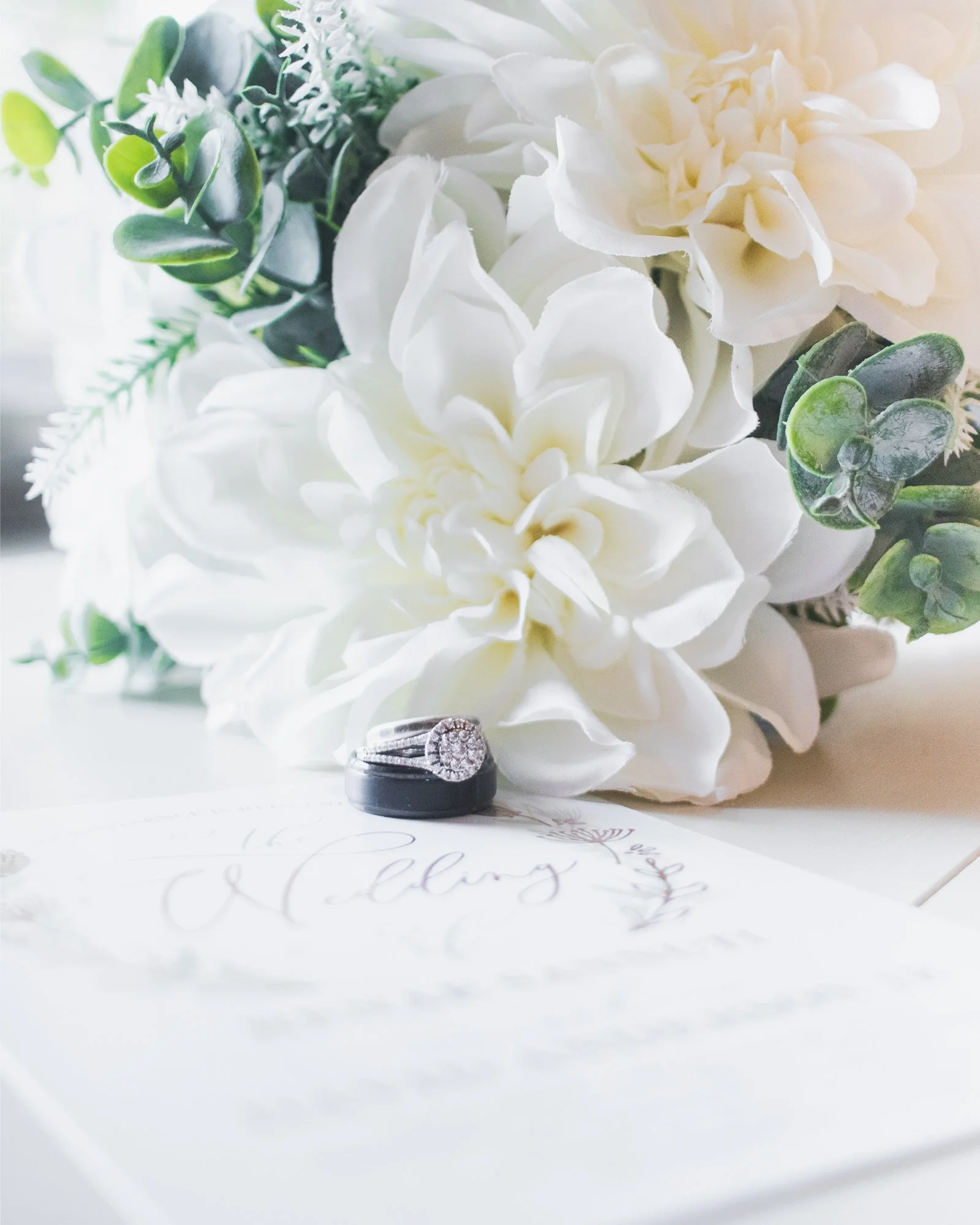 Wedding ring with diamonds resting on a white invitation card, with white flowers and green leaves in the background.