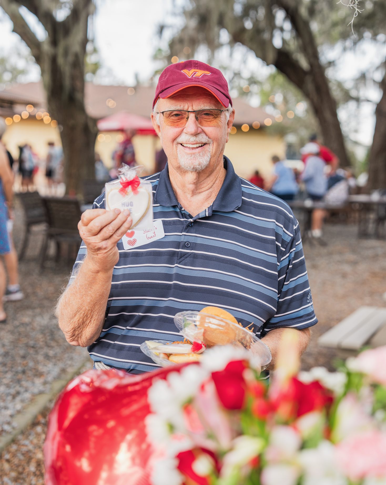 A man smiling at an outdoor event, holding a decorated cookie with the word 'HUG' on it and a plate of sandwiches. He is wearing a red cap with a Virginia Tech logo and glasses, with a crowd and string lights in the background.