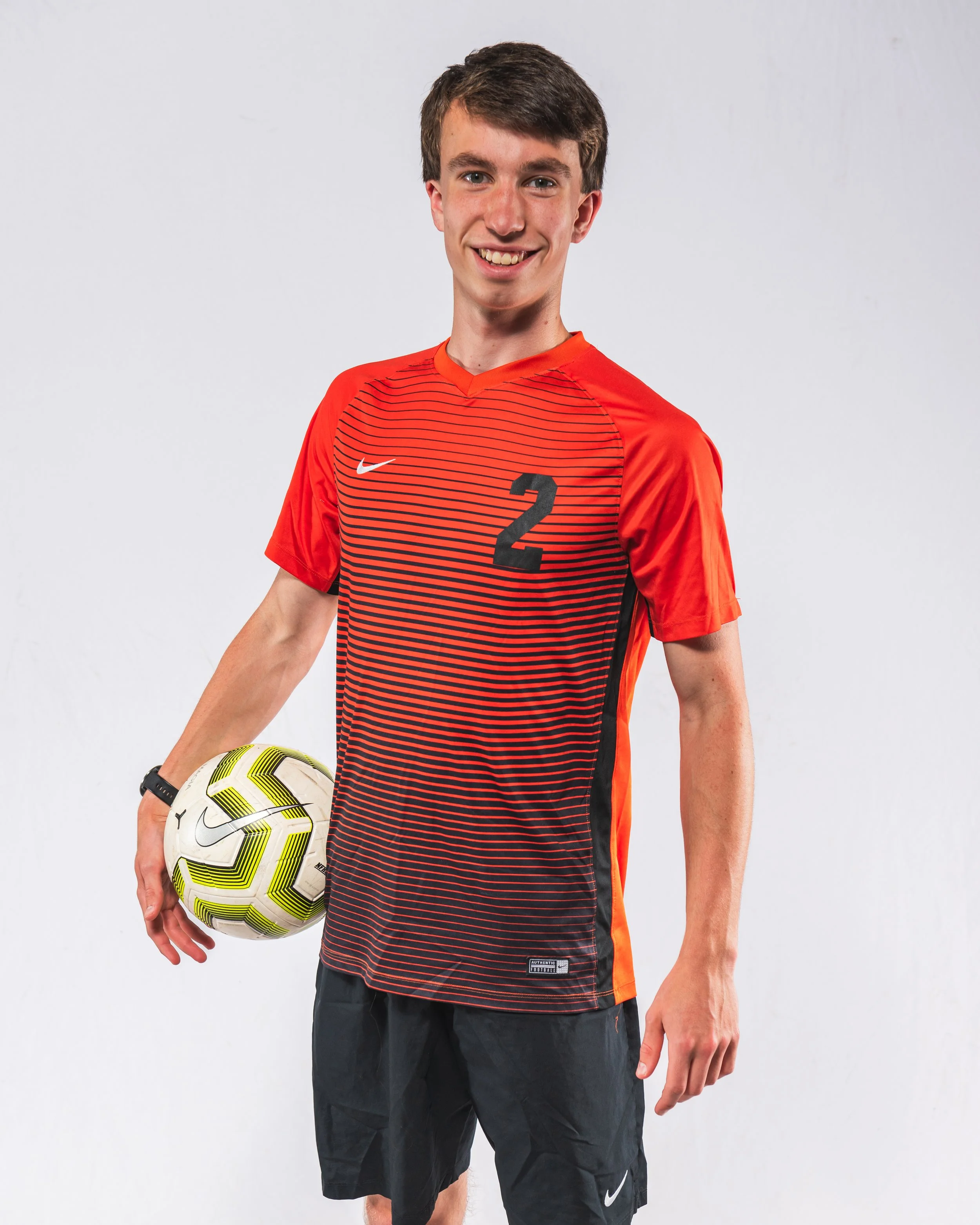 Young man in red and black soccer jersey holding a yellow and white soccer ball, smiling, standing against a plain white background.
