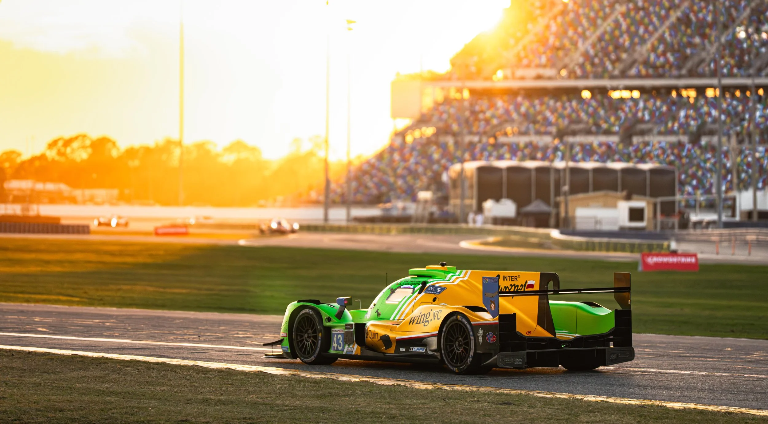 A race car with green and yellow livery on a race track at sunset, with grandstands filled with spectators in the background.