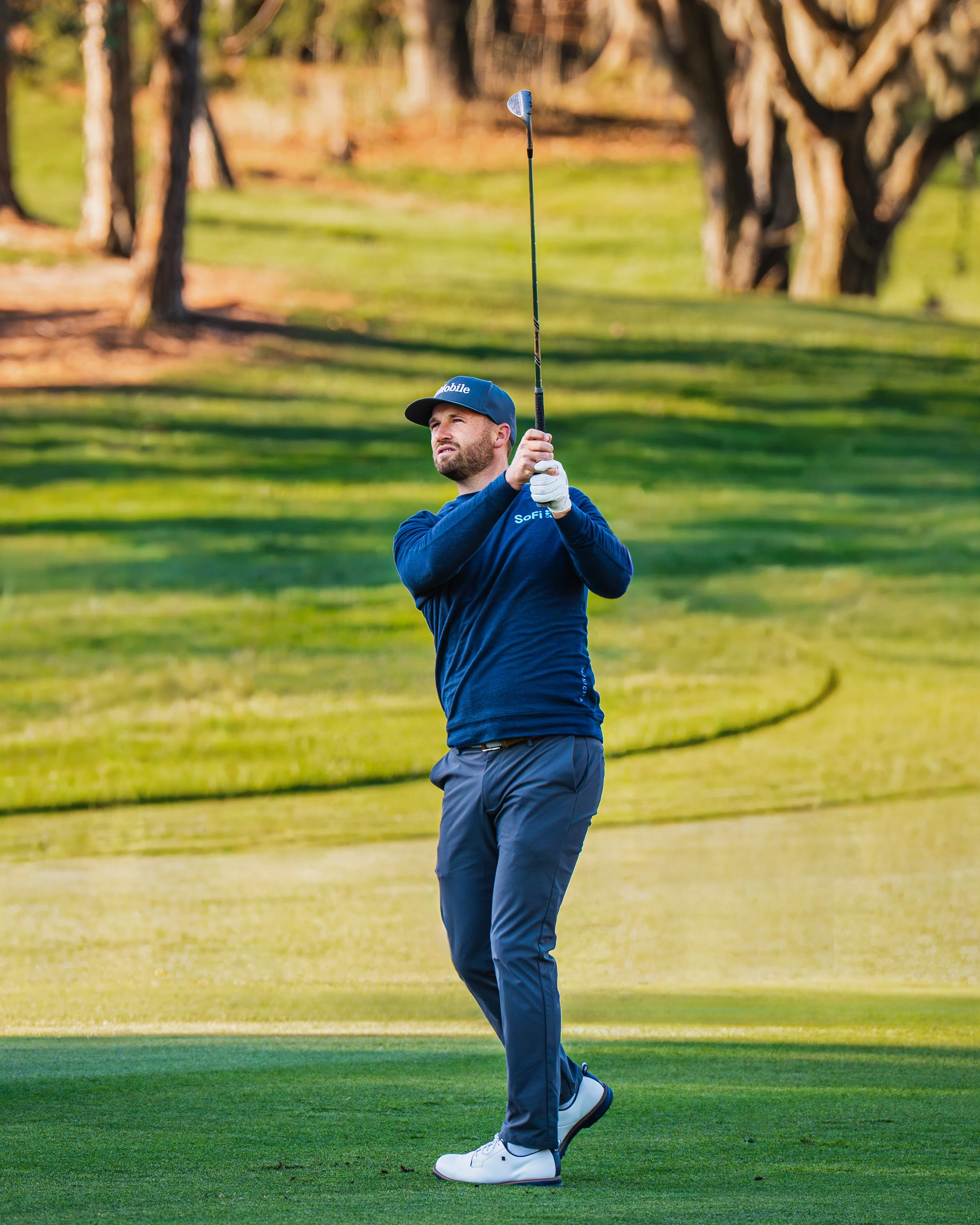 A man in a blue shirt and gray pants playing golf on a course, swinging a golf club.