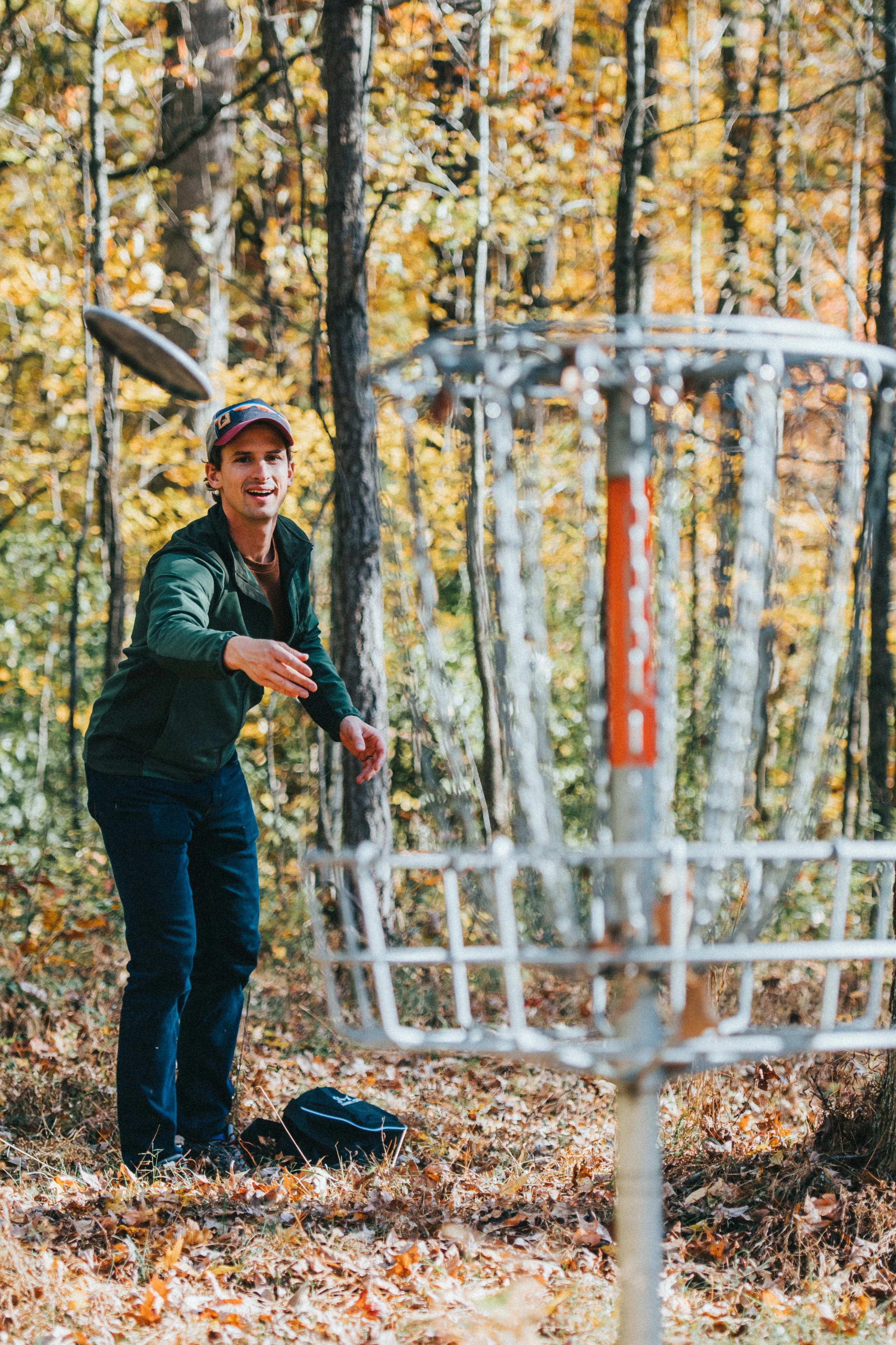 A man smiling in a forest during autumn plays disc golf, throwing a disc towards a metal basket.