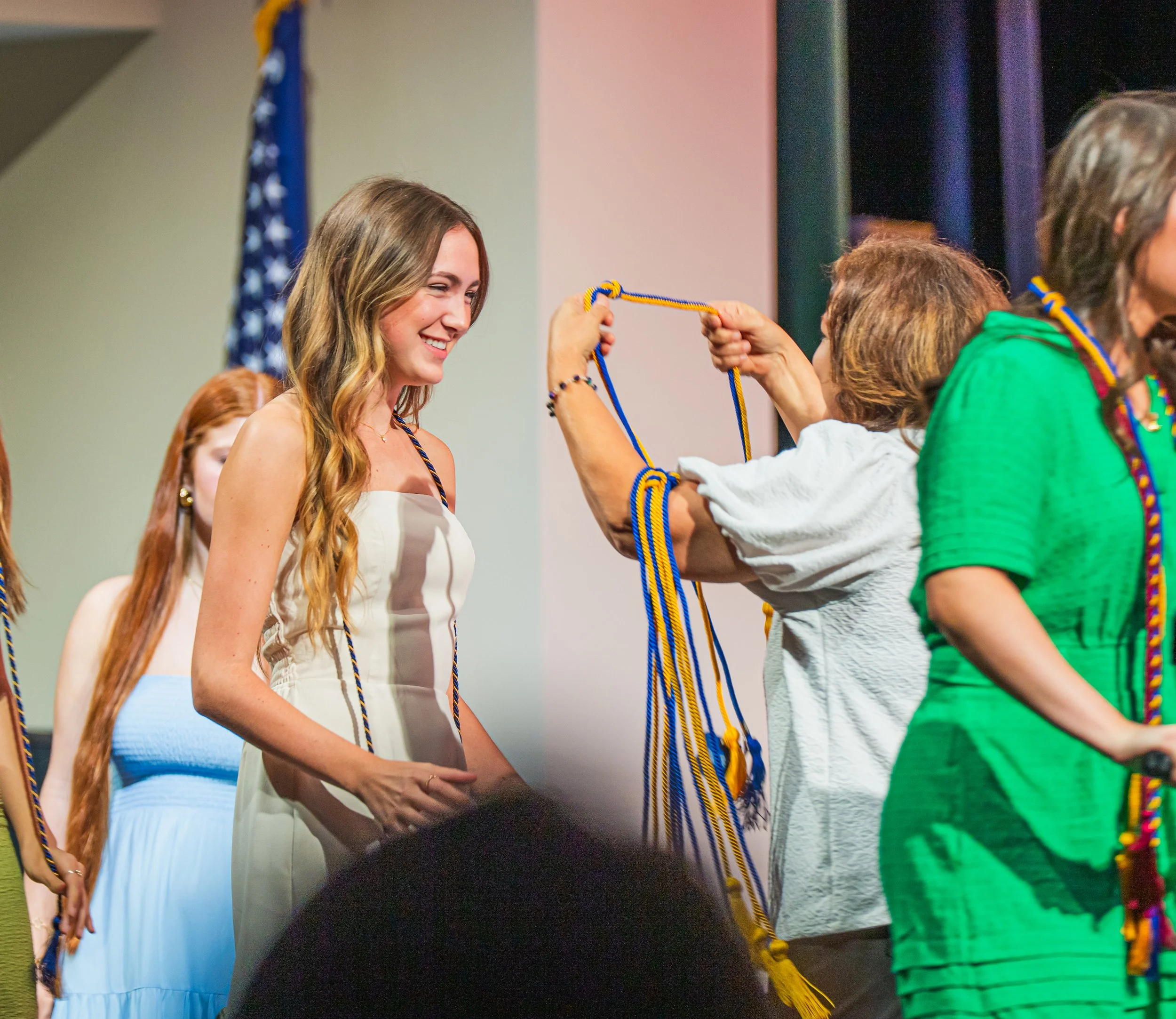 A woman with long wavy hair in a white dress is smiling as she receives a blue and yellow honor cord during a graduation ceremony, with other graduates and an American flag visible in the background. Graduation photos. Senior photography. 