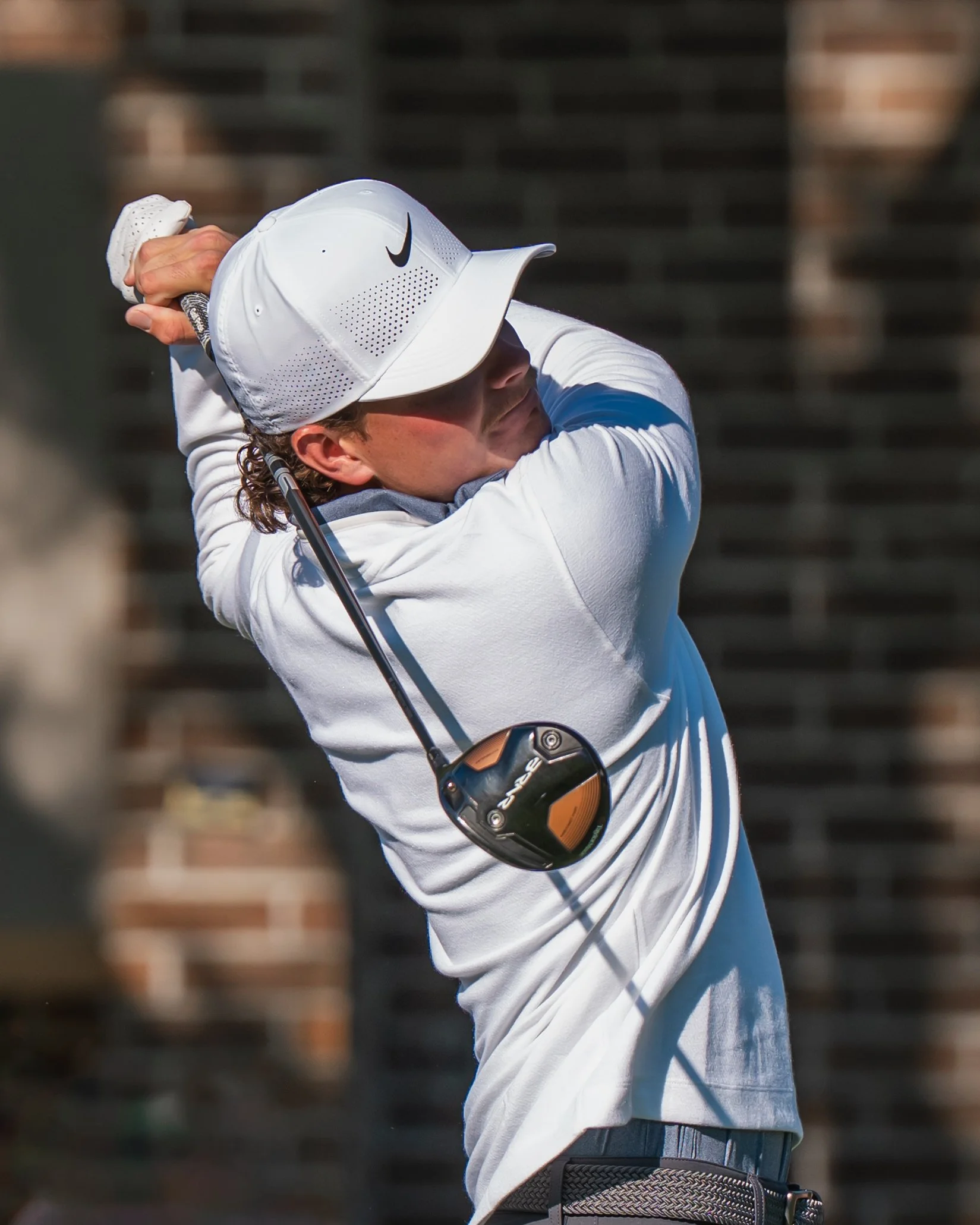 Luke Clanton, a golfer in a white Nike hat and white long-sleeve shirt swings a golf club, with a brick wall in the background.