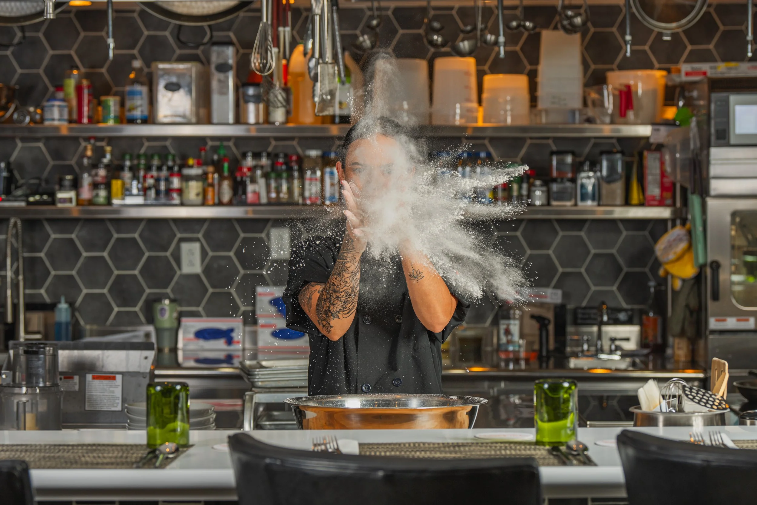 Portrait photography, Central Florida. Corporate headshots. A chef in a black uniform is throwing flour in the air behind a kitchen counter with utensils, glasses, and plates, in a restaurant or professional kitchen setting.