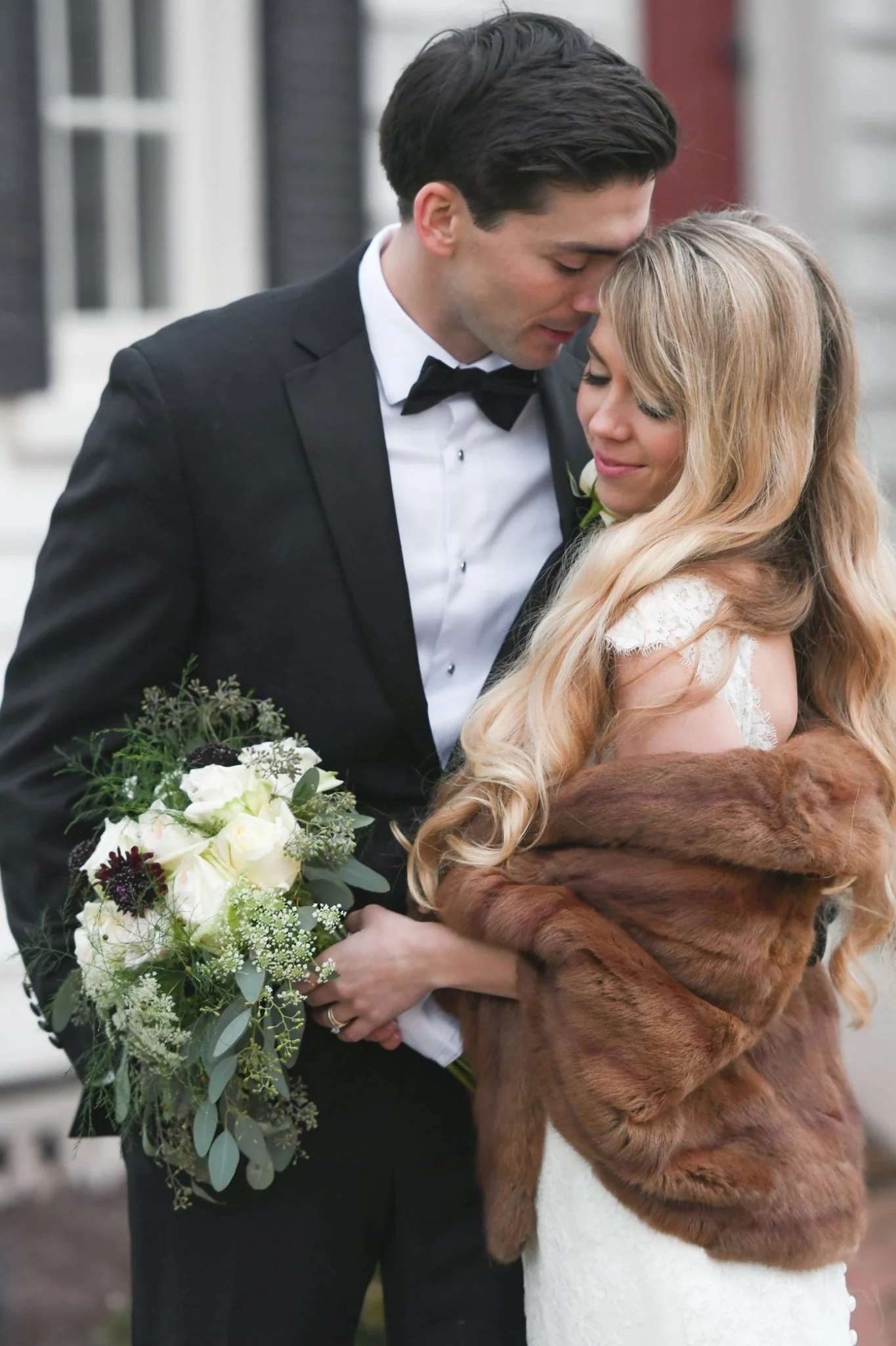 A bride and groom sharing an intimate moment, with the groom in a black tuxedo and the bride in a white wedding dress and a brown fur wrap, holding a bouquet of white and green flowers.